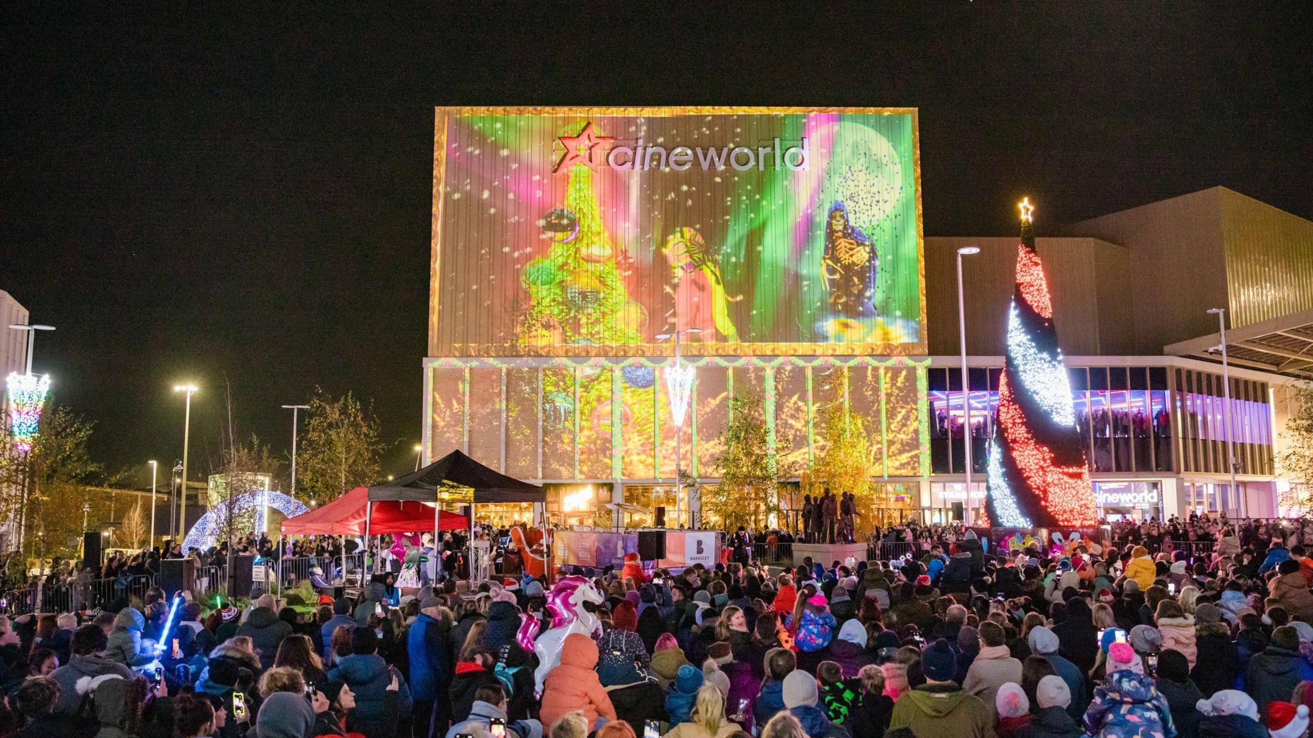 A picture of a town square, with several hundred people with their backs to the camera as they look up at a large animation projection covering the front wall of a cinema building.