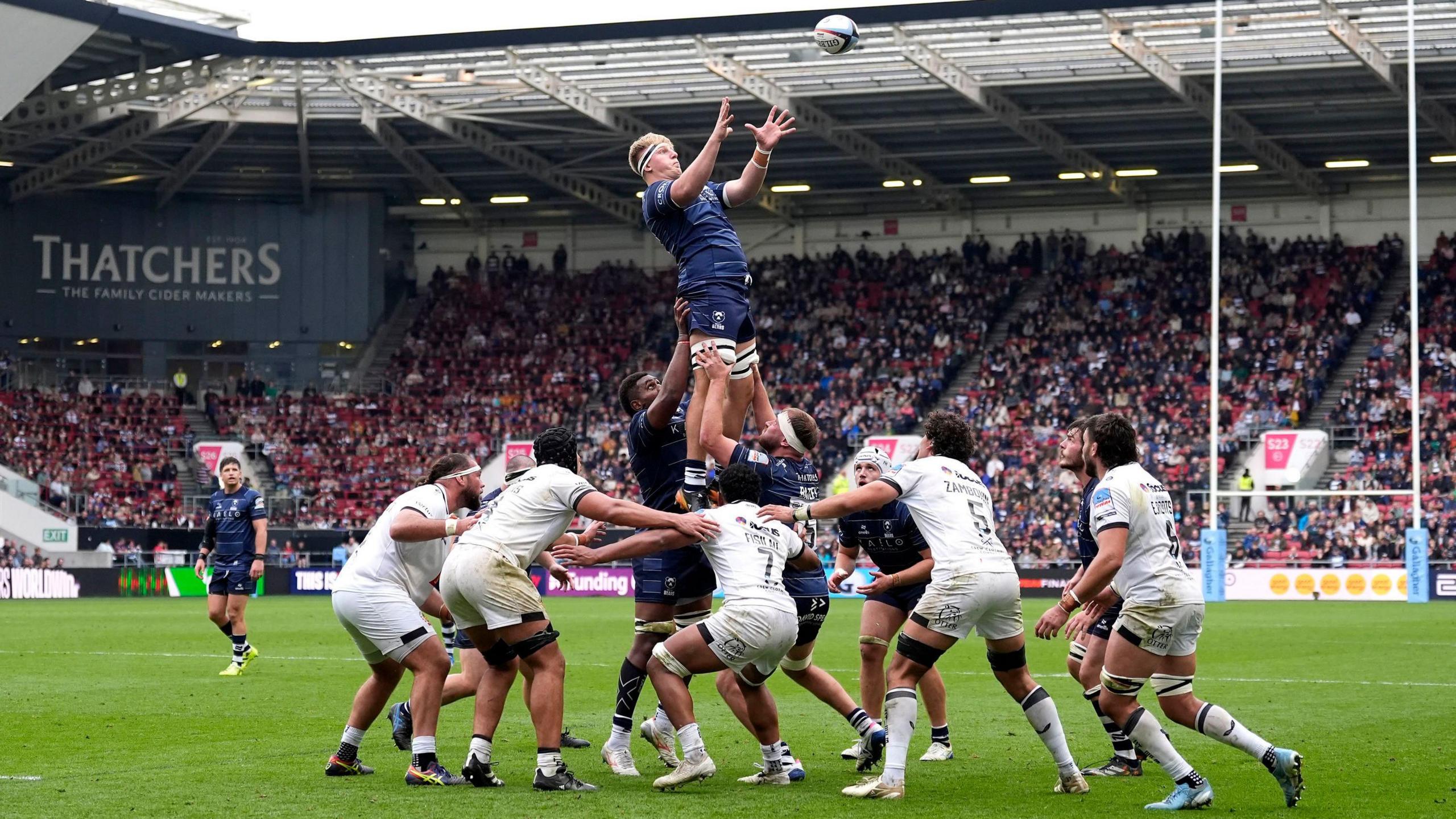 Bristol Bears' James Dun claims the ball from a lineout against Exeter Chiefs at Ashton Gate. He is being held up by his team-mates. Exeter players, in all-white kits, are contesting the line out. In the background full stands can be seen