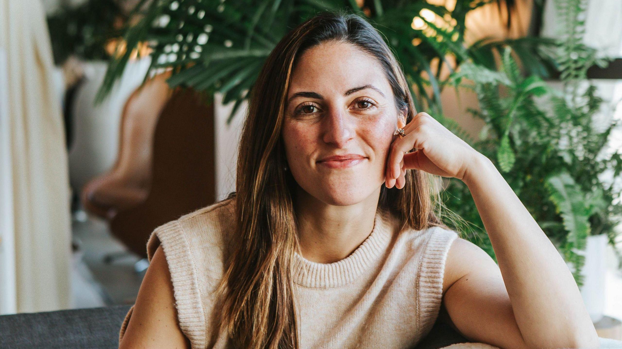 A woman with long brown hair is sat on a sofa smiling at the camera, with her hand touching her cheek. Wild houseplants are behind her.