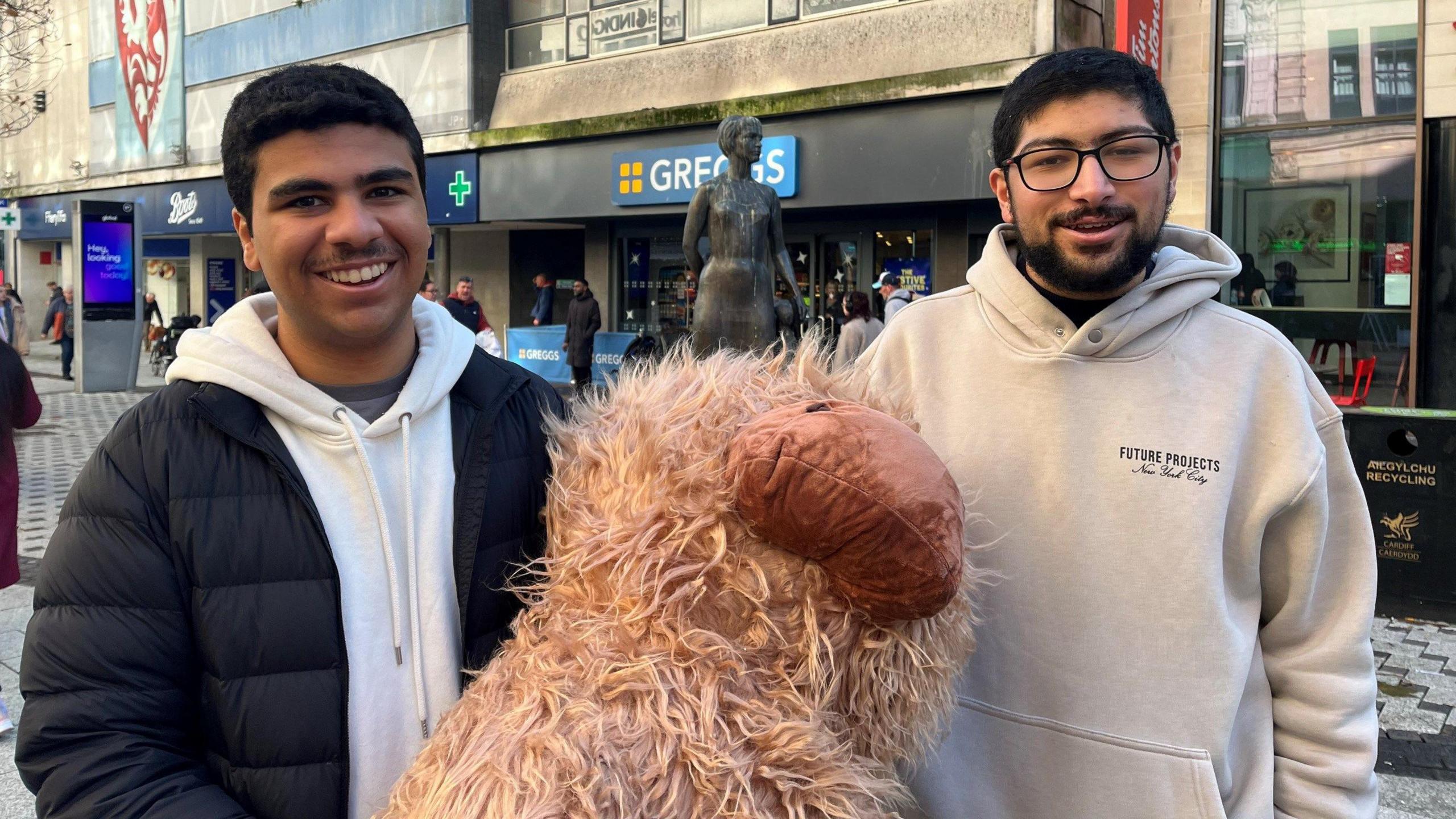 Students Hassan and Ali smiling at the camera holding a fluffy model of 
 a cow