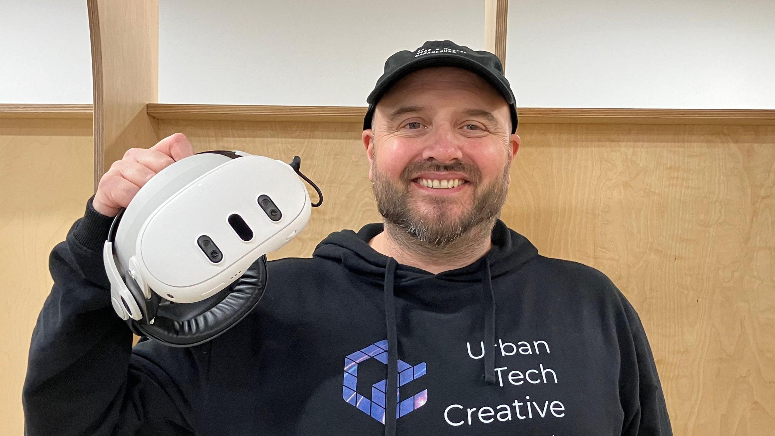 James Lee Burgess smiles at the camera while holding a white virtual reality headset up. He wears a black hoodie with his business name written on it.