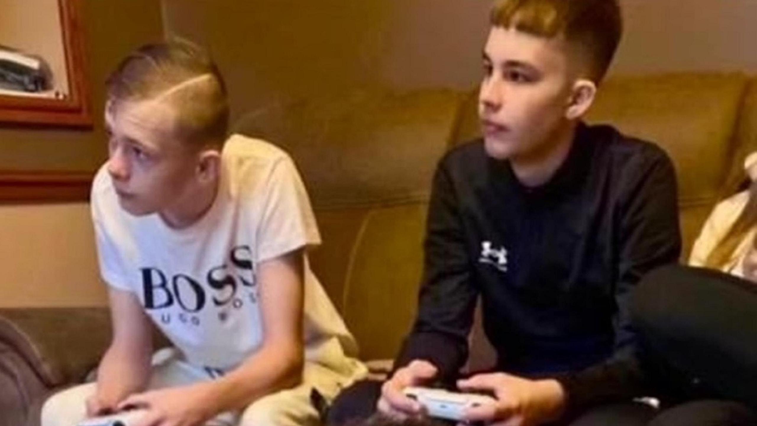 Two teenage boys sit on a sofa together, playing on a games console with controllers in their hands as they lean forward to focus