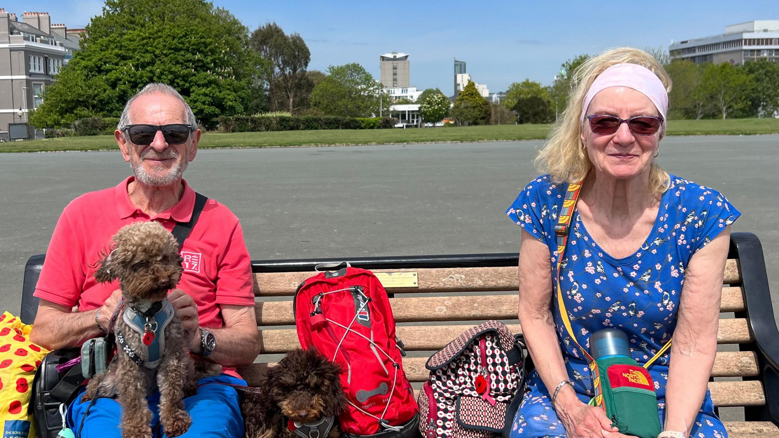 Bob and Rita Micek with their dogs Chico and Rolo sat on a bench on Plymouth Hoe. Rita is wearing a blue floral dress, and Bob is wearing a red t-shirt with blue shorts. One dog is sat on his lap, while the other one is sat beside him.