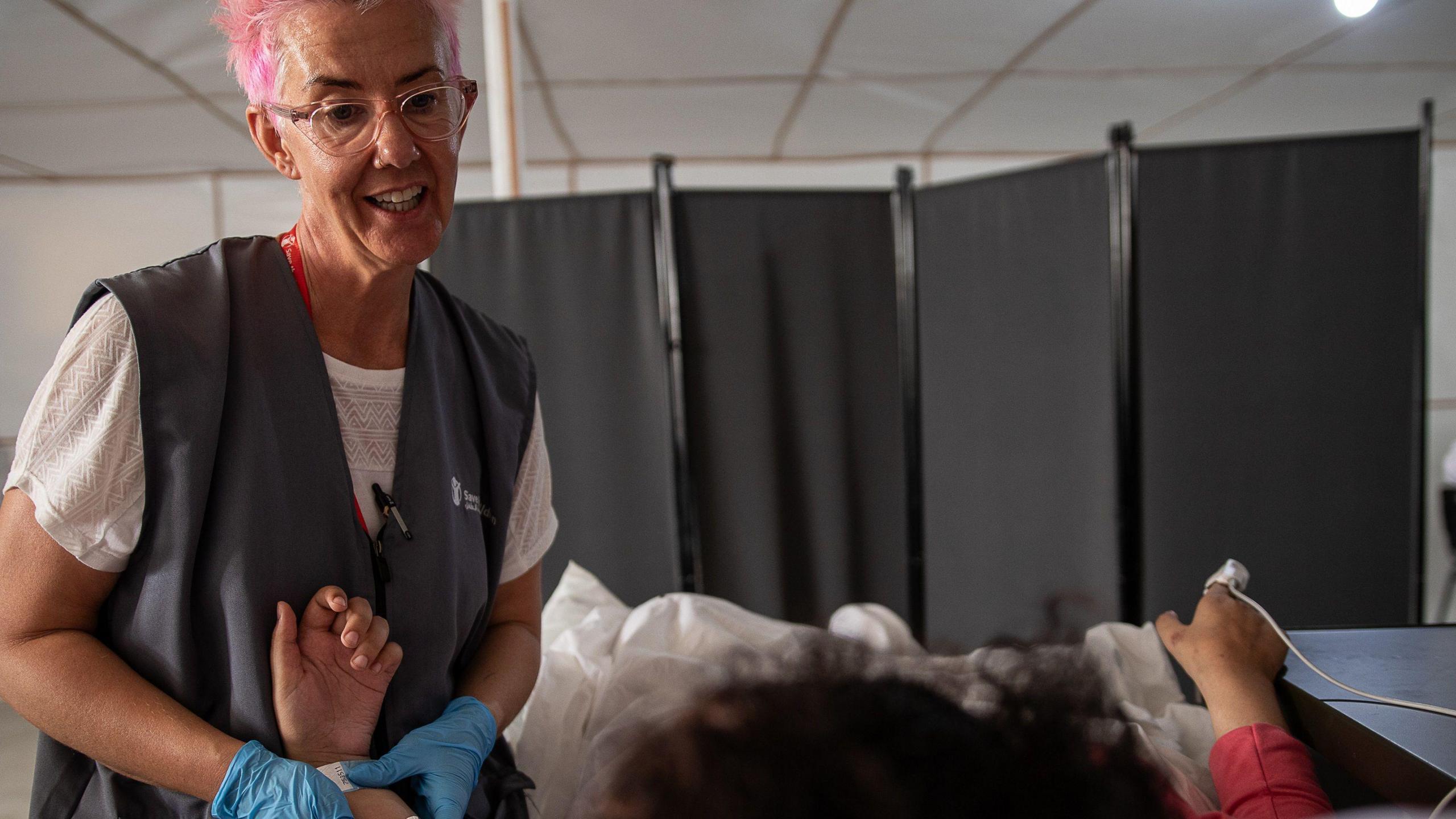 Nurse Becky Platt, with pink hair, is smiling as she treats a child patient whose face we cannot see