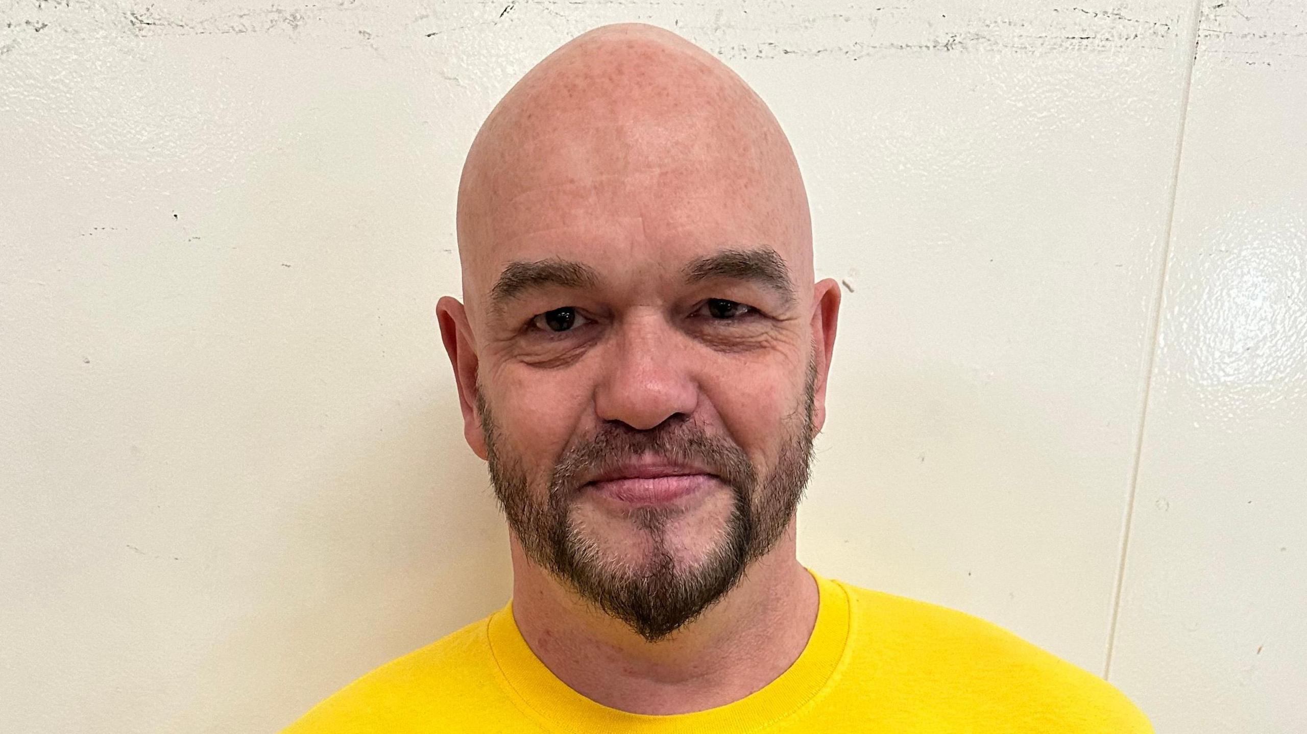 A bald man with a beard wearing a yellow shirt looking at the camera.