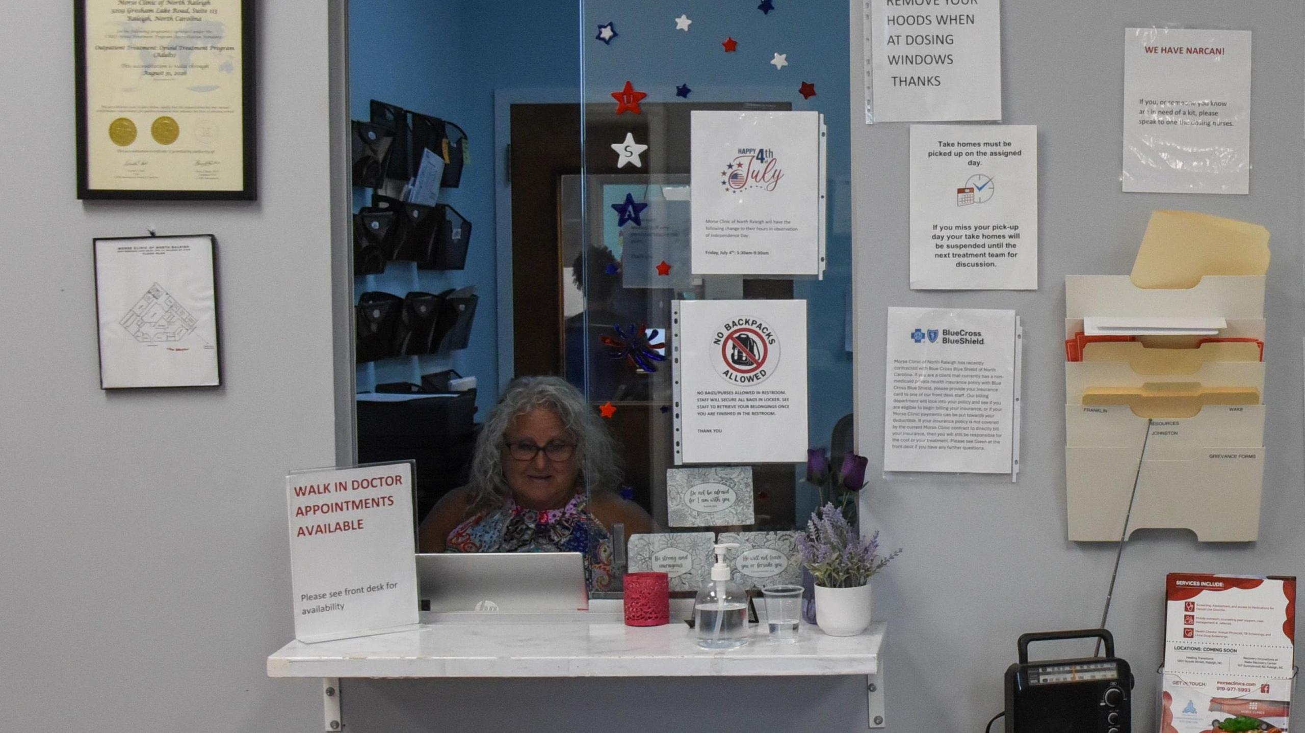 A worker sits behind a reception window at a clinic. Printed notices adorn the surrounding wall