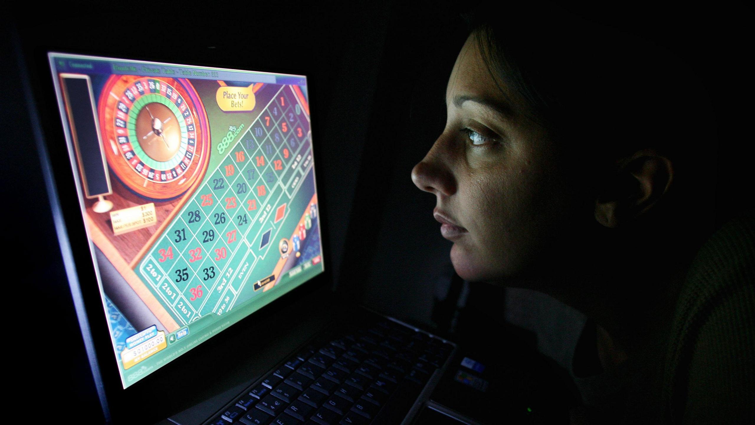 A woman plays roulette online in the dark. She is staring at her computer screen with just the light of the screen illuminating her face.