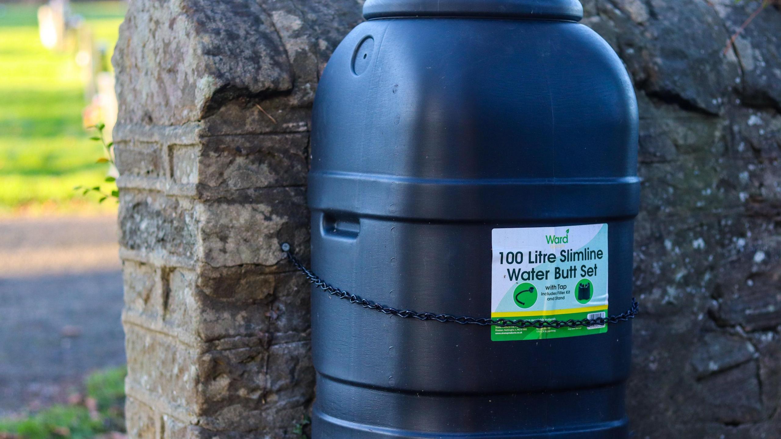 A black water butt stands next to an old wall