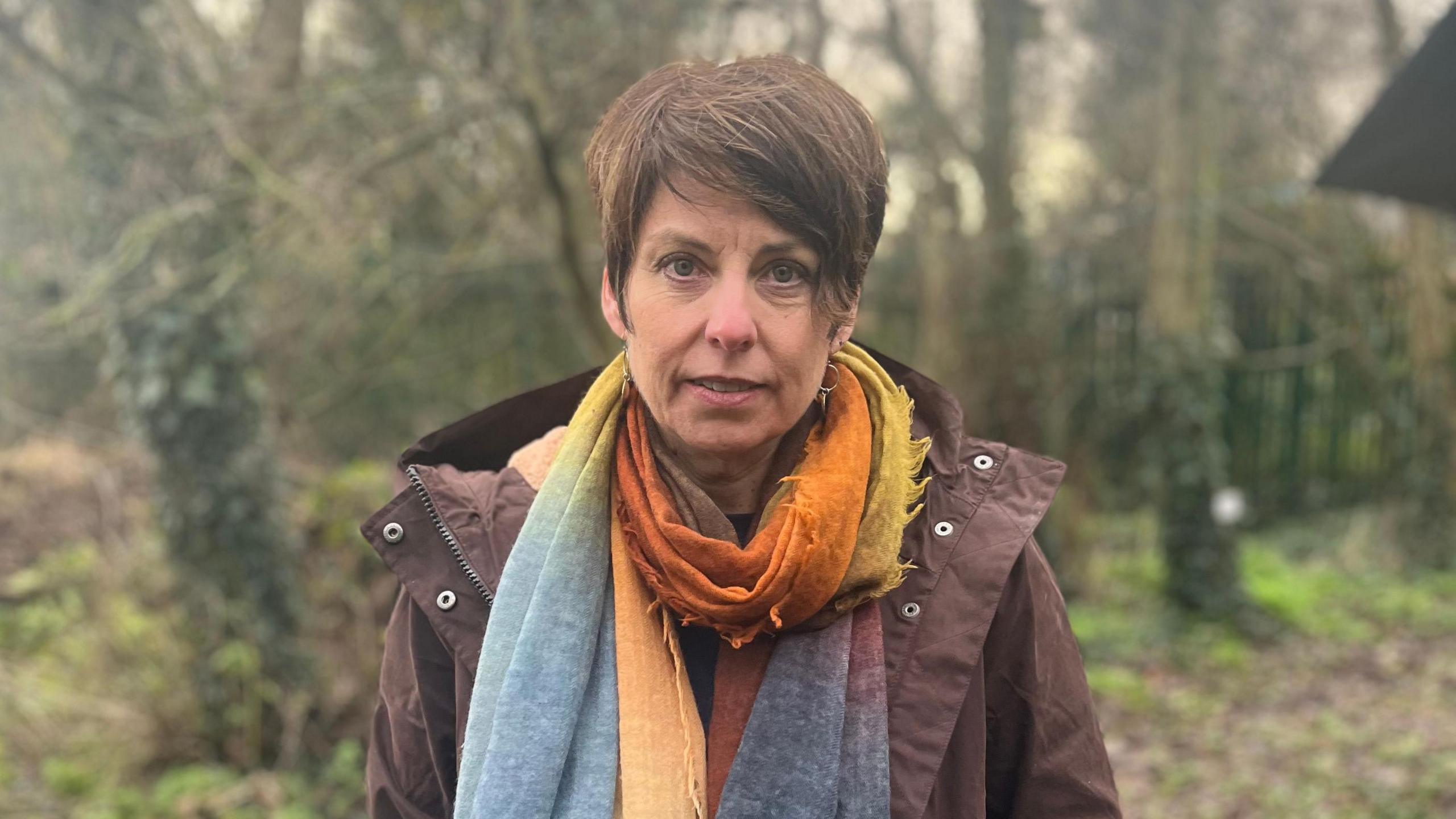 Lisa Morrison, a woman with short, brown hair, looks directly into the camera with a neutral expression.  She is standing in a wooded area, wearing a brown hooded jacket and multicoloured scarf. 