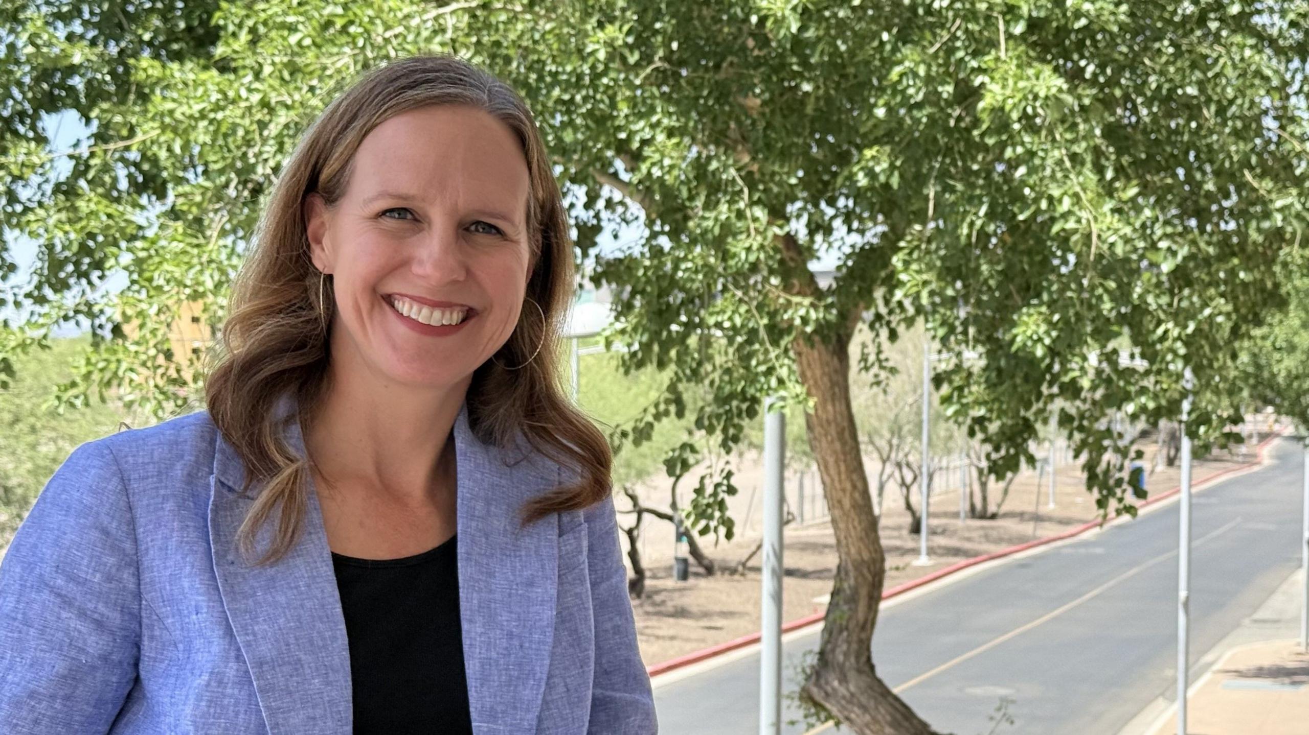 Sarah Skidd, in a blue jacket, smiles with a tree in the background.