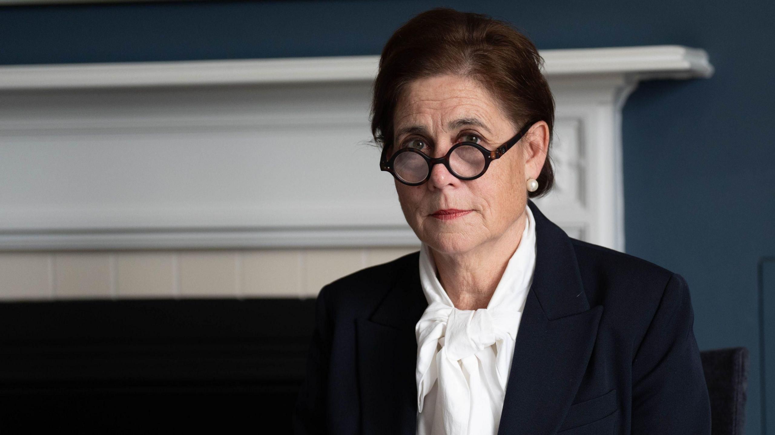 Baroness Lampard sits in the inquiry office at Arundel House in London. She is wearing a white shirt and navy suit. Her glasses are rounded and tortoiseshell in colour and she is looking direct at the camera with her hair up. She is wearing pearl earrings.