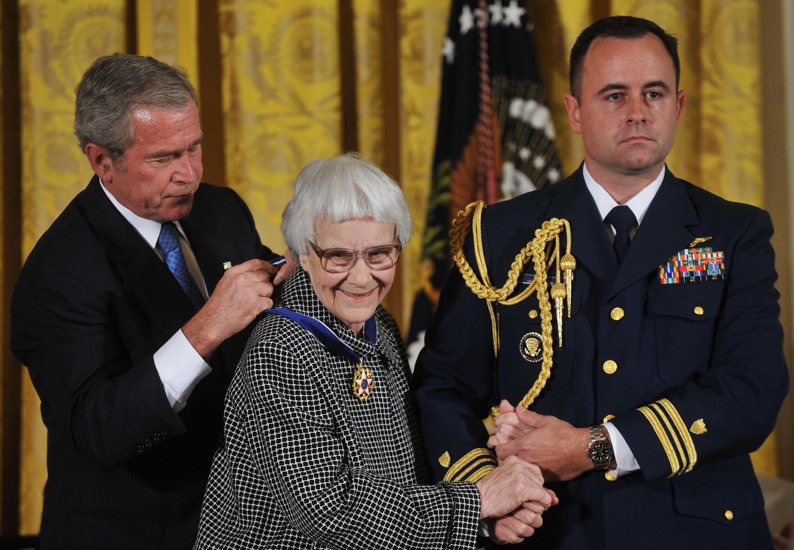 President George W. Bush in dark suit and blue tie, putting the Presidential Medal of Freedom around Harper Lee's neck. Harper smiling, wearing glasses, dressed in black and white dog tooth outfit, holding hand of man in uniform