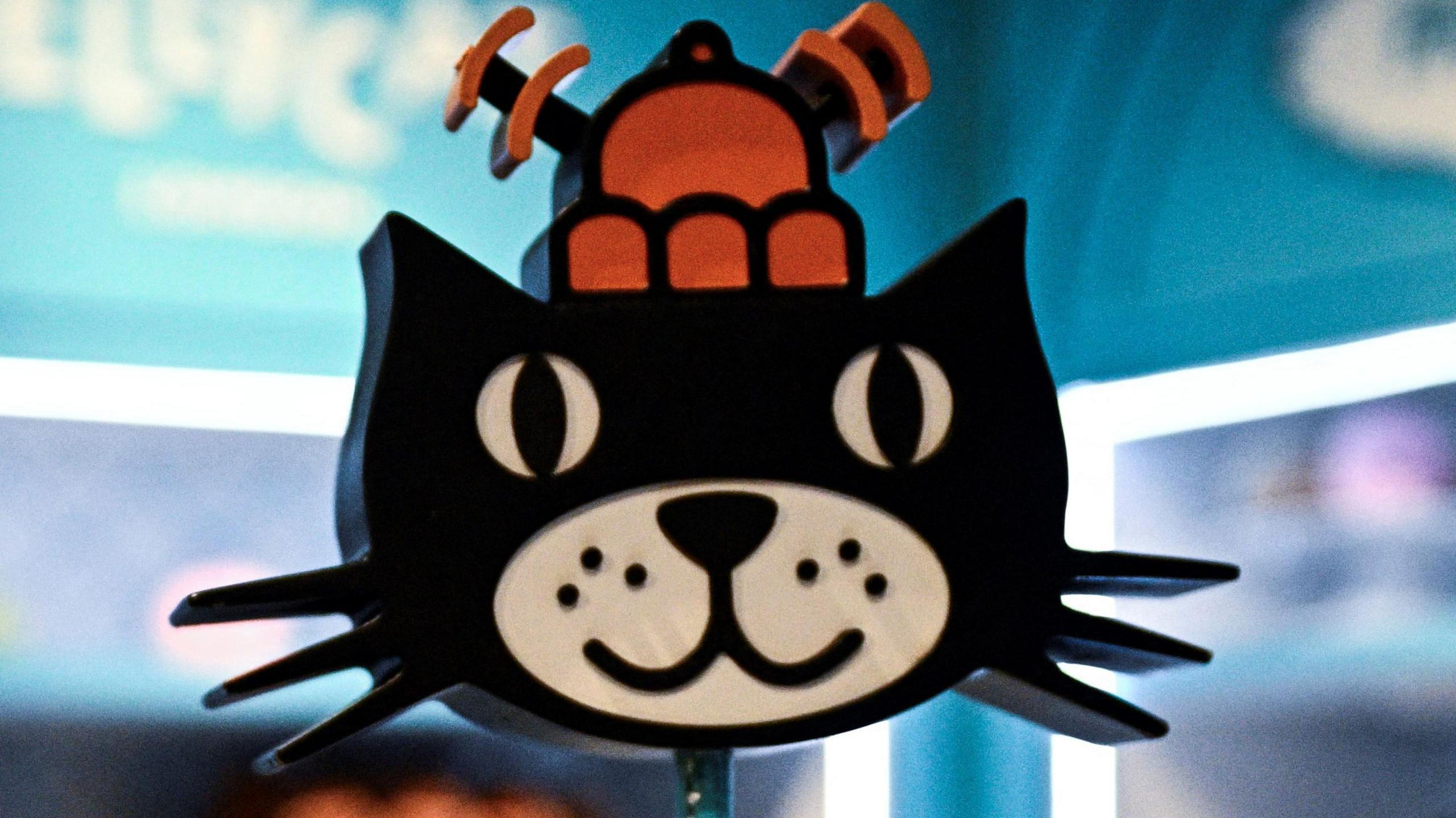 A image of the Jellycat logo. It shows an animated black cat with an orange hat on its head. The cat is smiling.