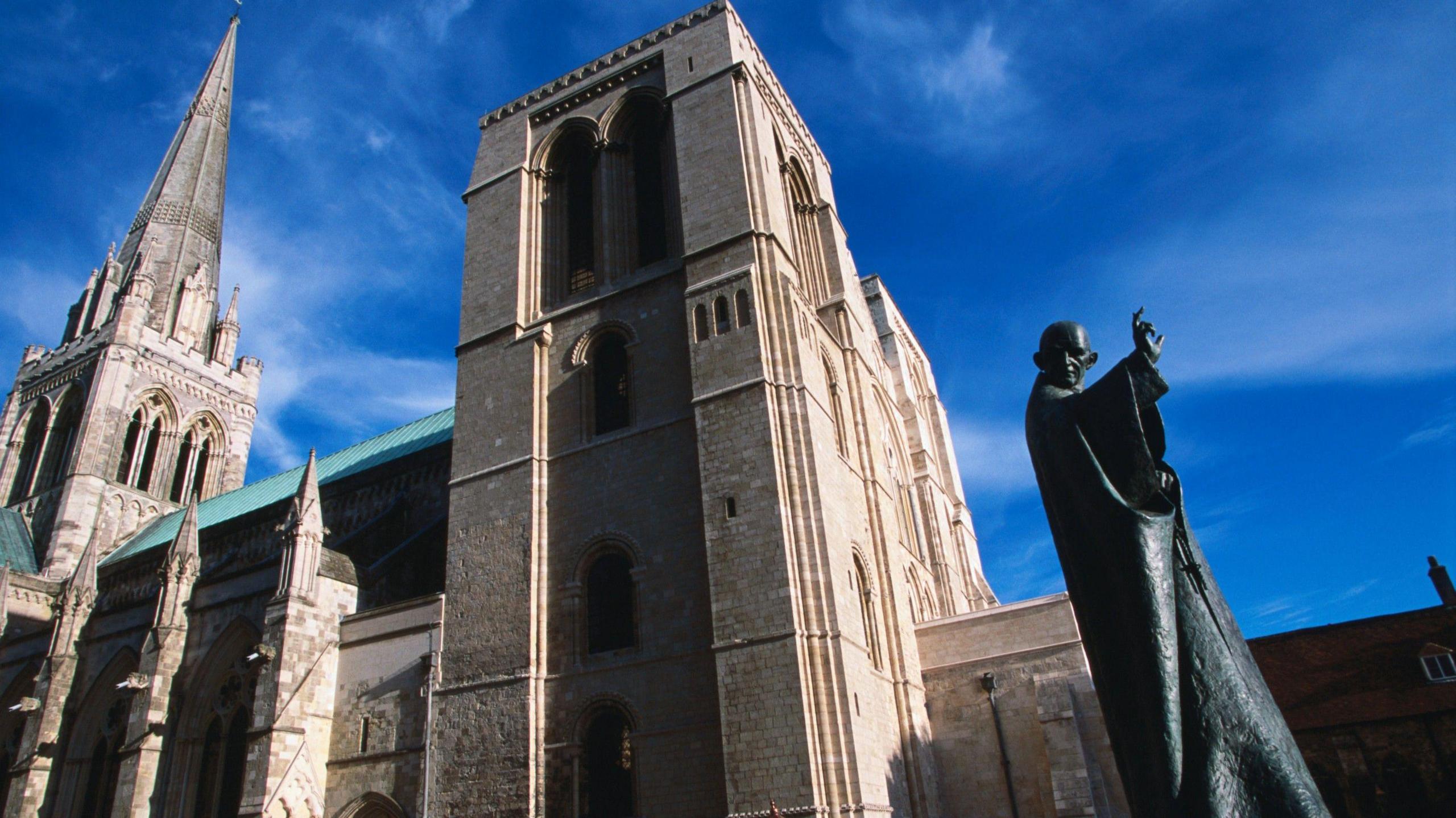 The exterior of Chichester Cathedral, with the grey Gothic building set against a bright blue sky and the modern statue of Saint Richard in the foreground.