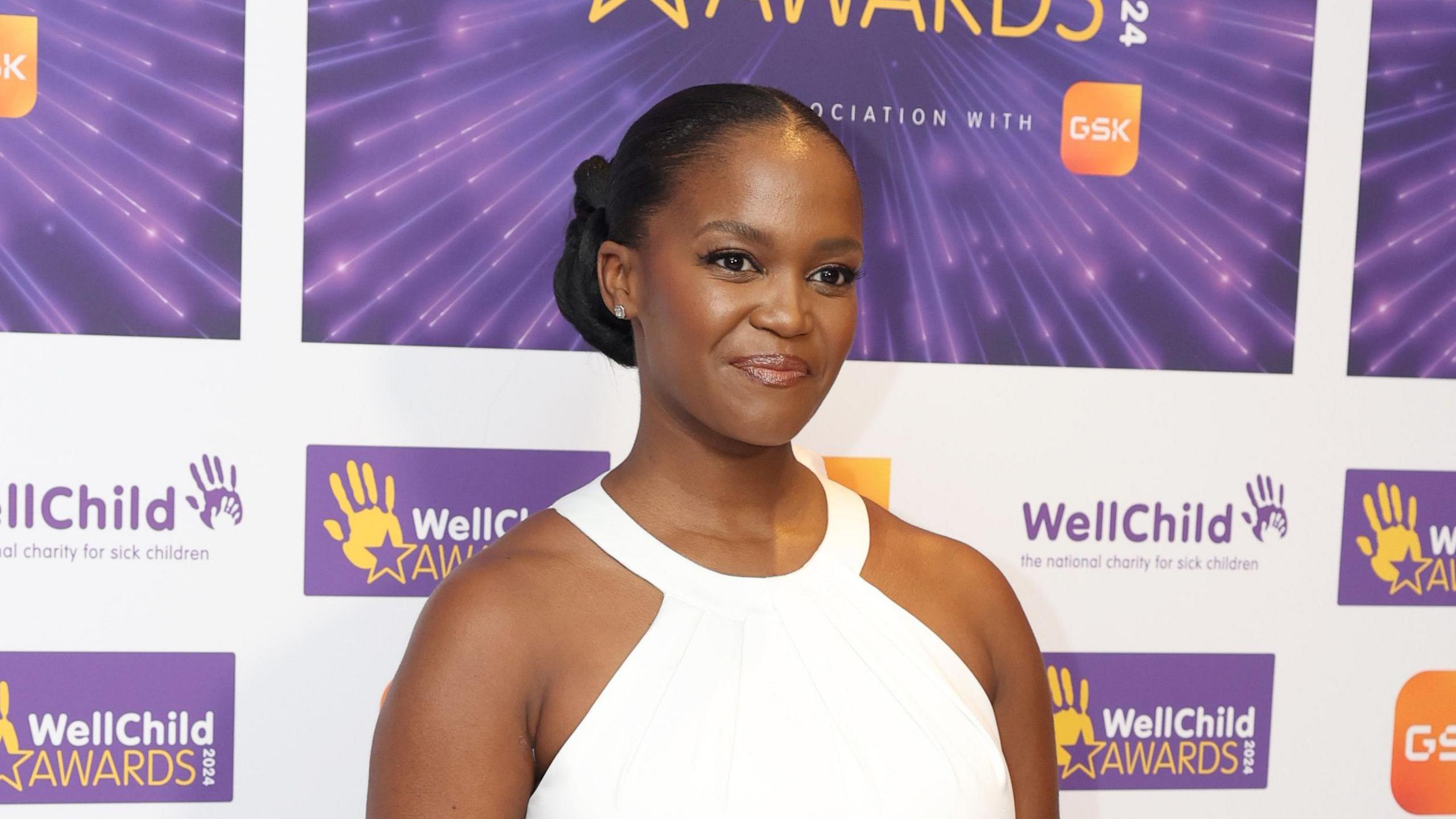 Oti Mabuse wearing a white dress standing in front of backdrop promoting WellChild Awards