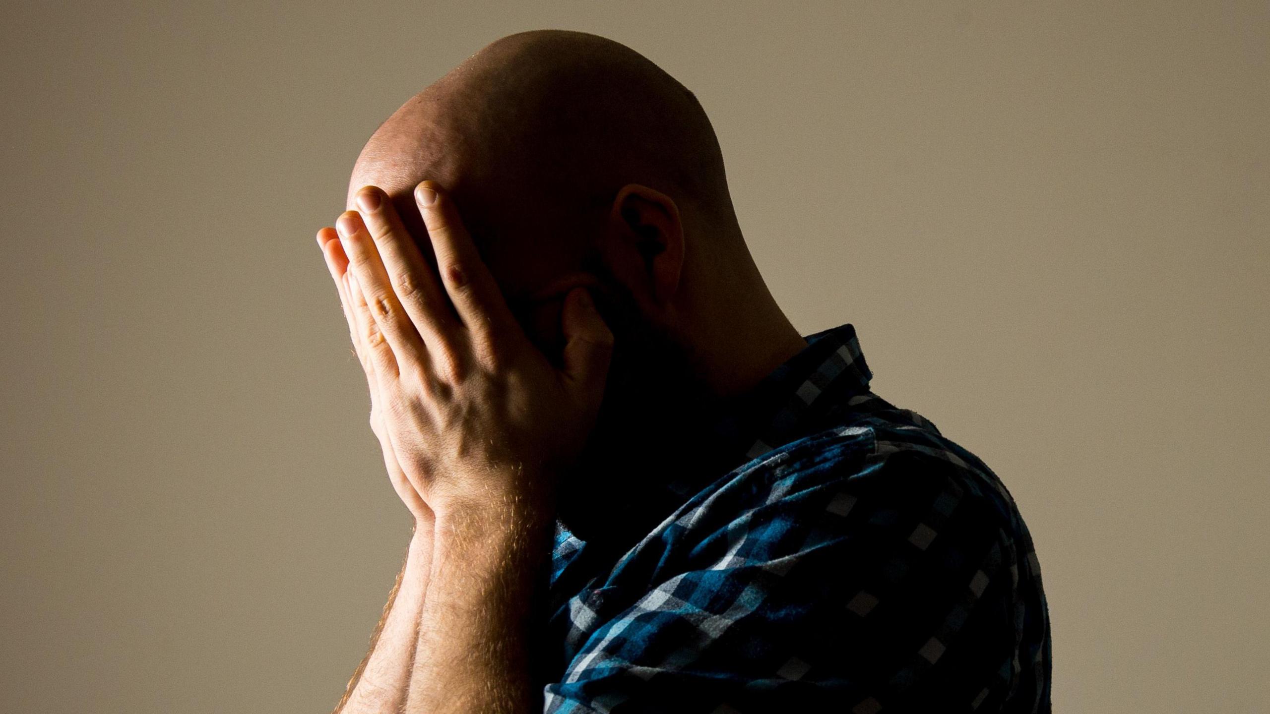 A man wearing a blue checkered shirt and holding his head in his hands.