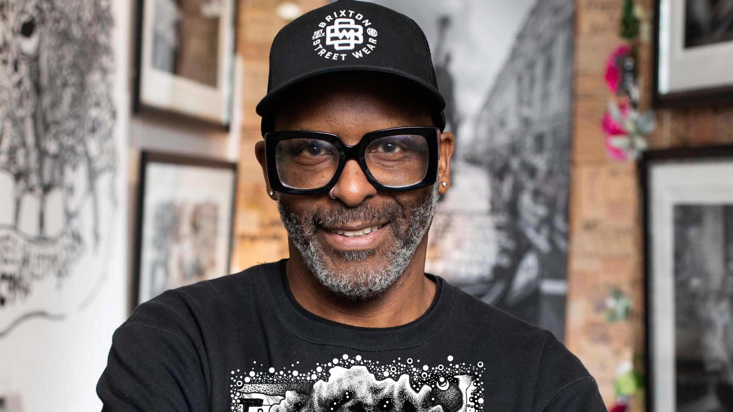 Head and shoulders image of Wayne Campbell in a black t-shirt and thick black glasses. He is also wearing a black cap saying Brixton Street Wear on it and his smiling. He has a silver nose ring. The background is blurred.