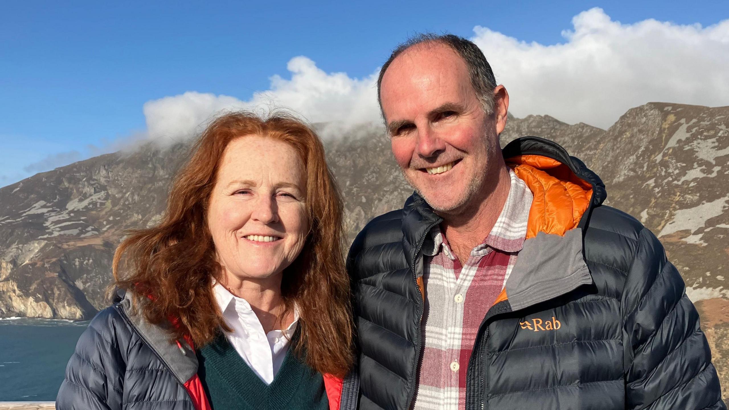 A man and a woman are staring into the camera. The woman has long red hair and is smiling. She is wearing a white shirt, dark v-neck jumper and navy anorak. The man has receding dark hair and is wearing a check shirt and dark anorak. Slieve League is in the background of the picture and the sky and water are also visible.