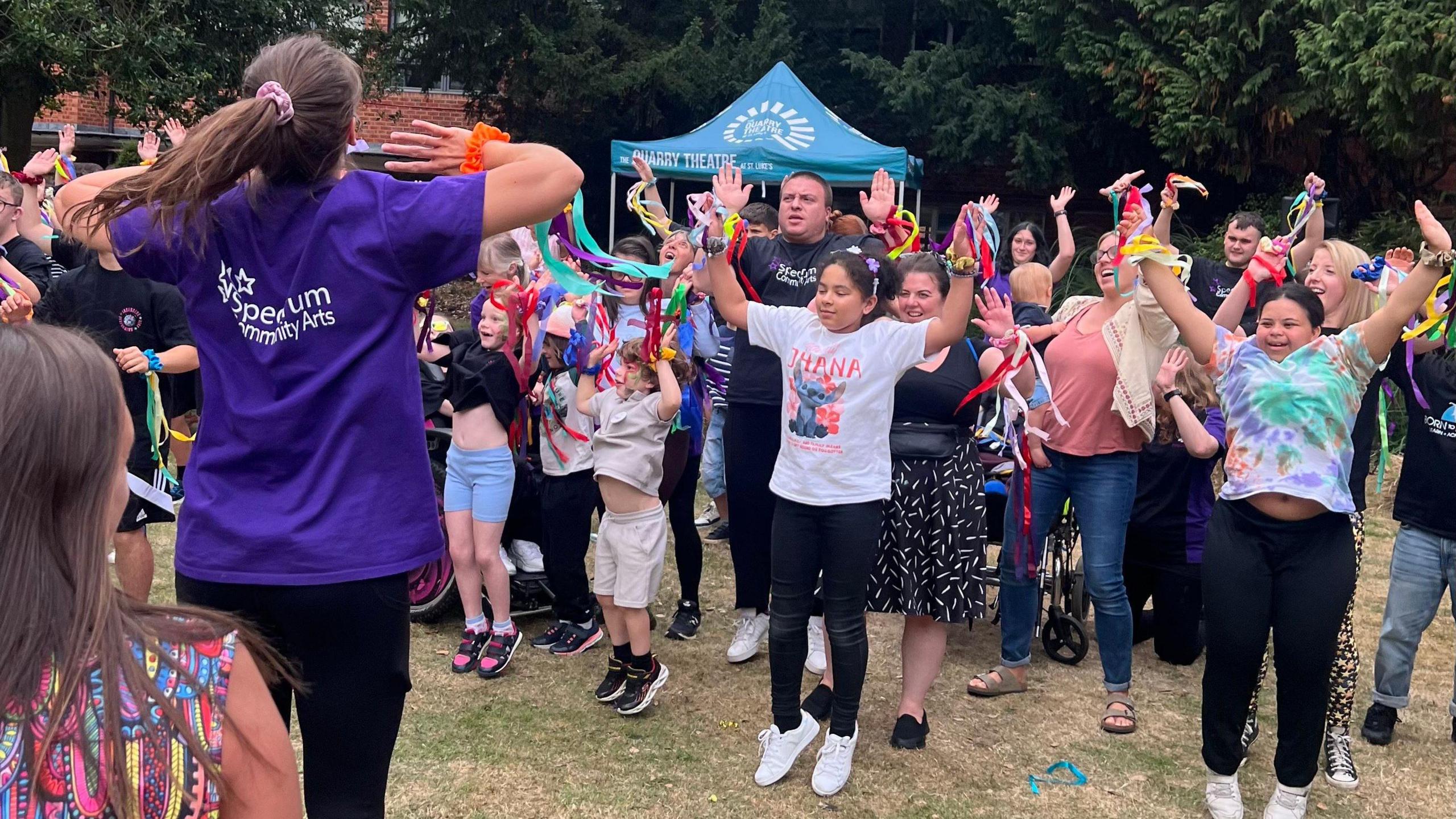 A large group of people taking part in a dance event - jumping up in the air. A woman is wearing a purple T-shirt. There is a marquee distance. They are smiling and laughing. 