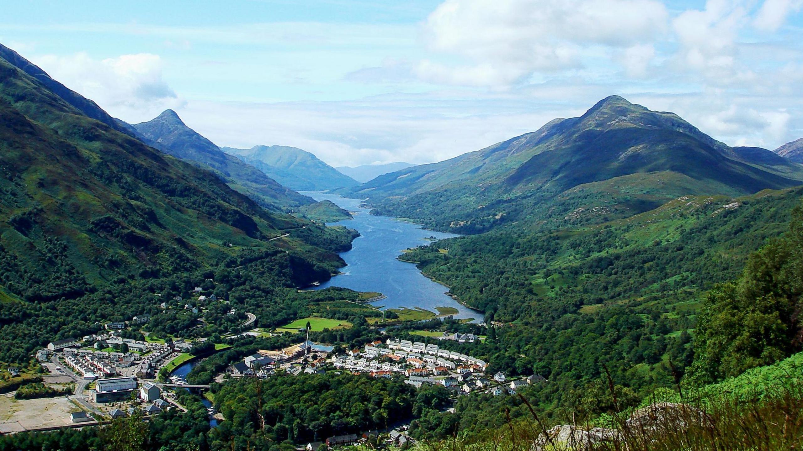 The village of Kinlocleven is nestled in a heavily wooded glen by the shores of a loch. The village is surrounded by hills and mountains.