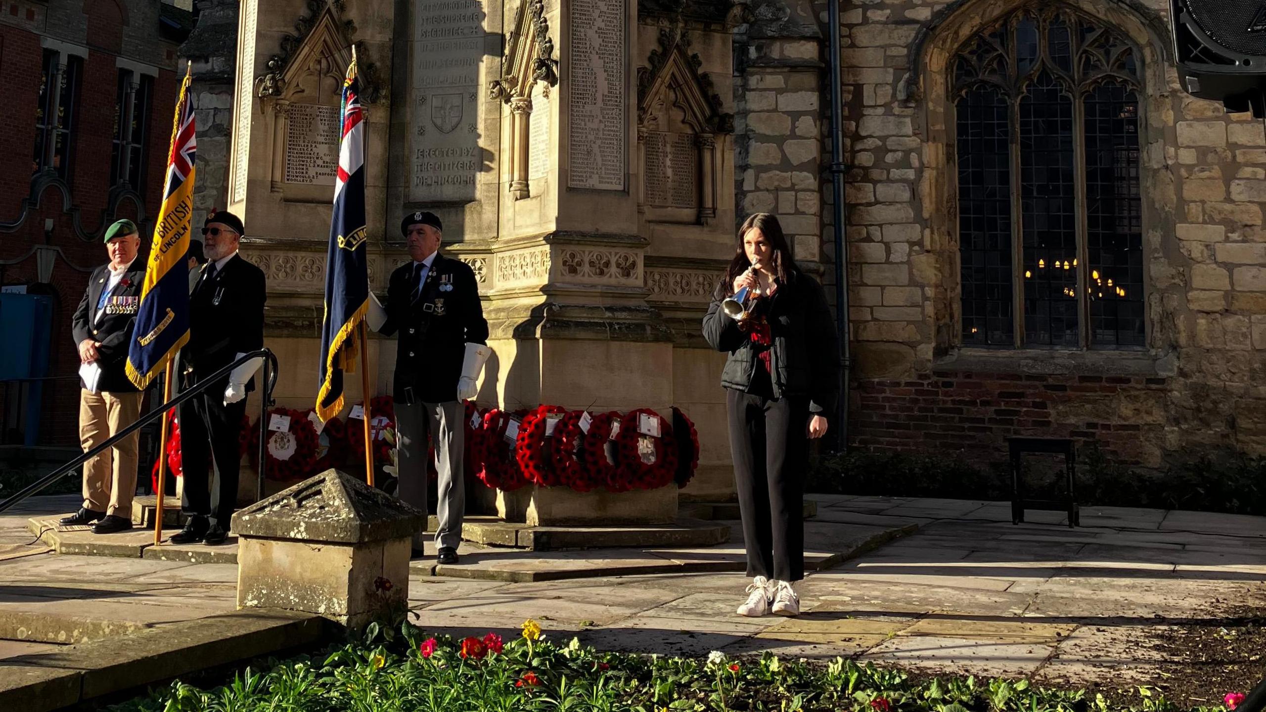 Three men in blazers with medals and regimental caps hold flags outside a church and war memorial as a young woman dressed in black plays a trumpet. The war memorial is adorned with poppy wreaths.