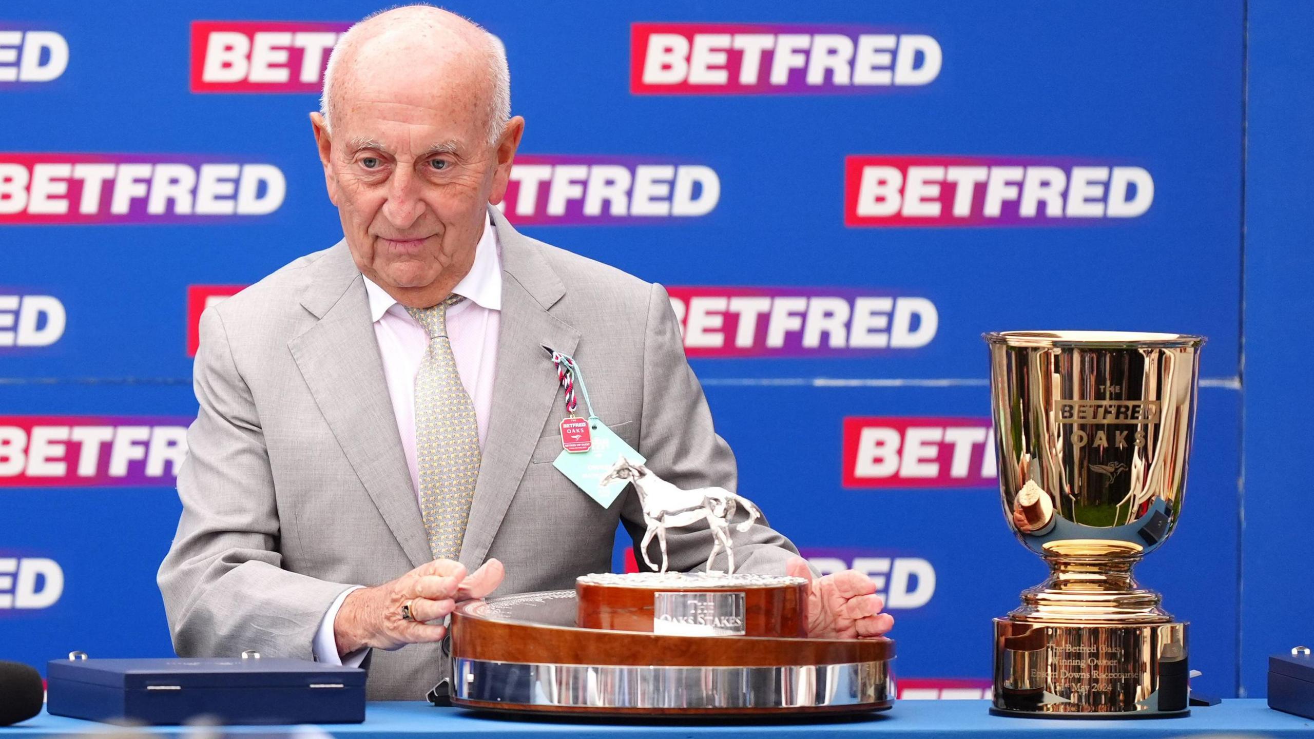 Fred Done, wearing a grey suit, grey tie and white shirt, prepares to present the trophy, which is wood and silver with a small silver horse sculpture on top, on Ladies Day at the BetFred Derby Festival, while standing alongside a gold goblet-shaped trophy and in front of a red and blue BetFred advertising