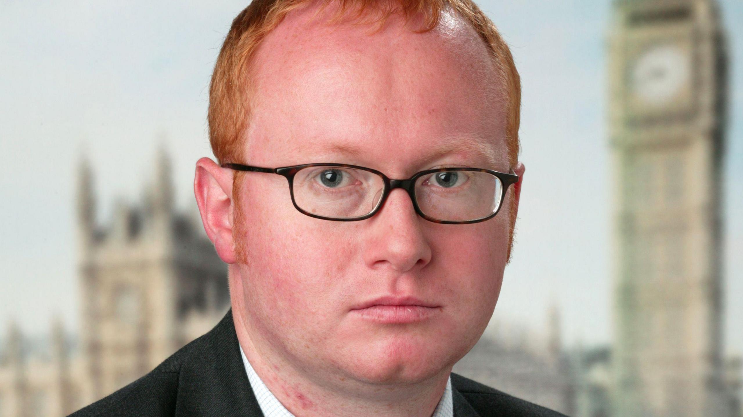 Luke Akehurst. The picture is a close-up of Akehurst's face. He has short red hair and is wearing dark-rimmed glasses. The Houses of Parliament can be seen out of focus behind him.