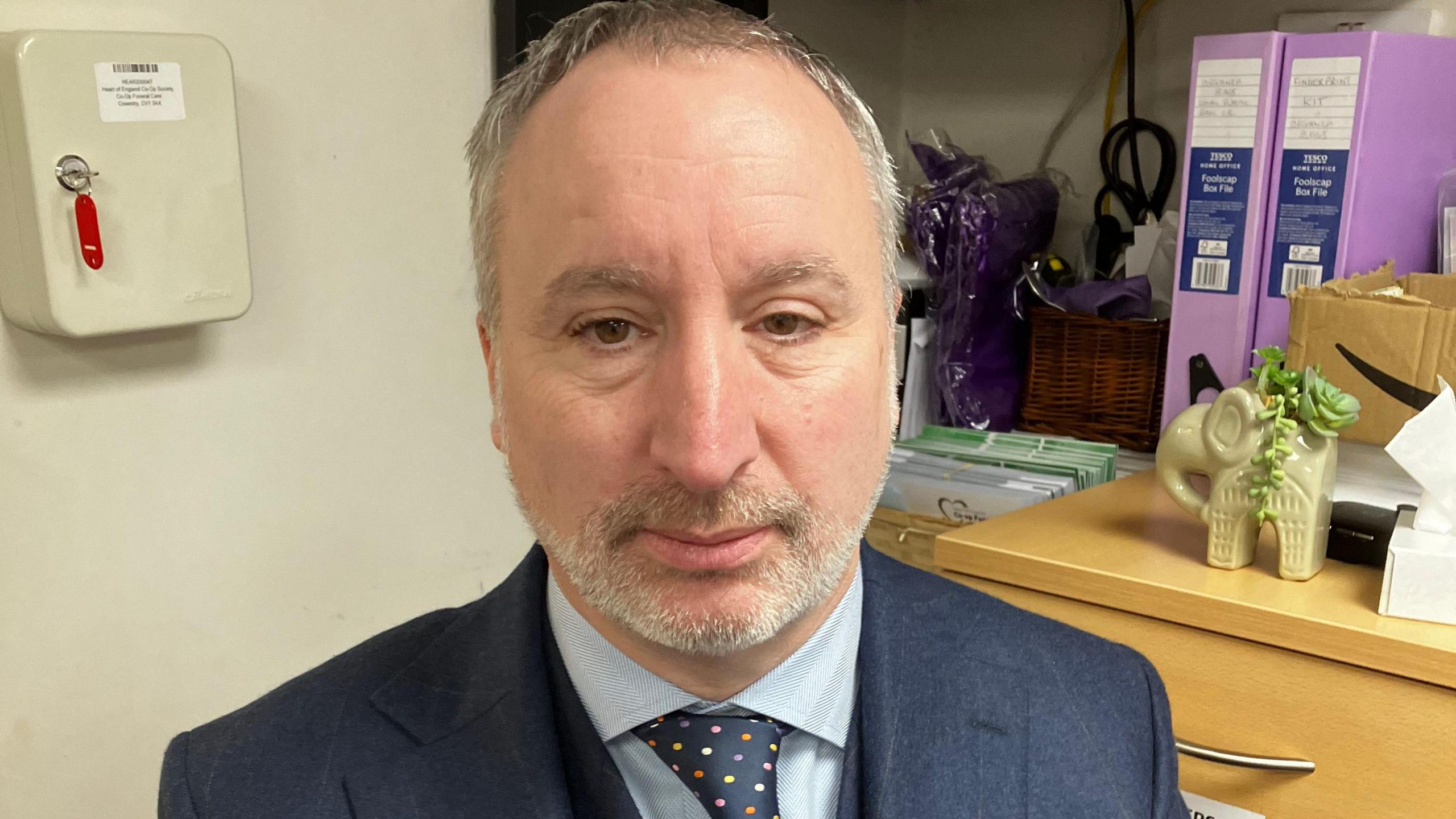 A man with neatly trimmed salt-and-pepper hair and white beard in an office. He is wearing a navy suite, blue shirt and navy spotted tie. Oak drawers, a wall safe and purple files can be seen in the background.