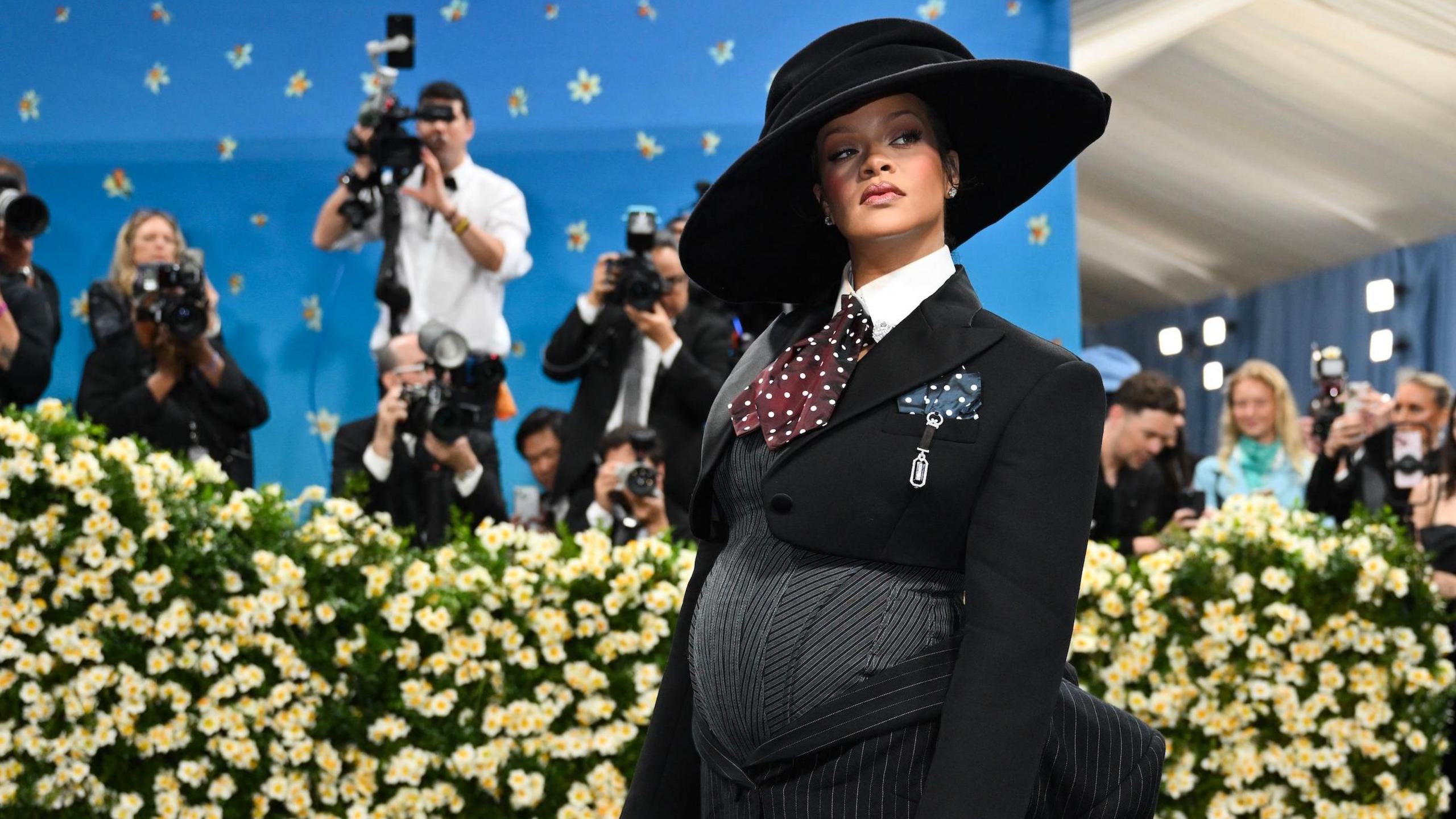 Rihanna appears at the Met Gala wearing an outfit tailored to her pregnant stomach. Photographer stand behind a bank of daffodils to snap pictures of her.