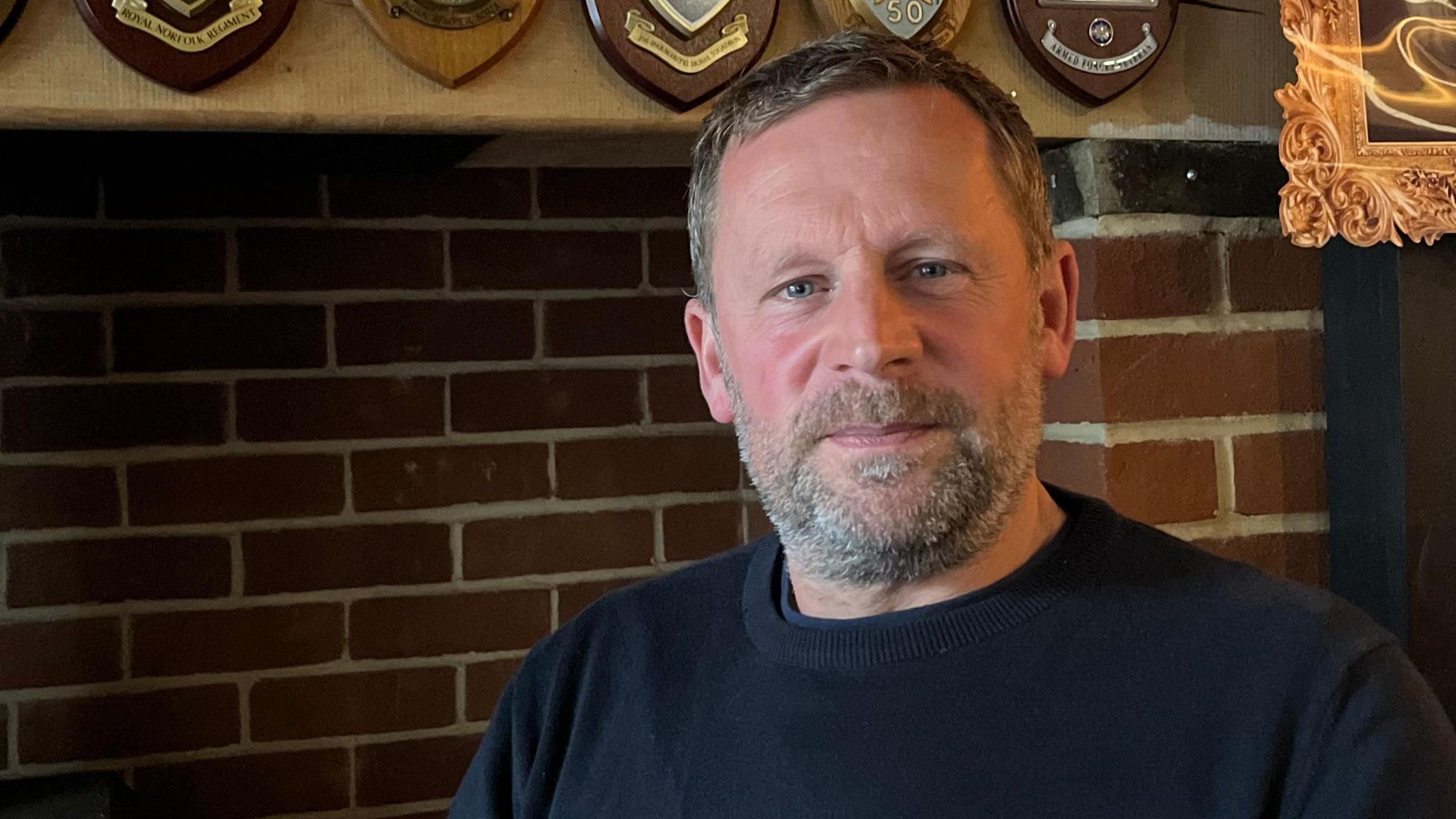 Nick Anema has a serious expression as he poses for the camera. He is seated before a red-brick fireplace and is wearing a dark round-neck jumper. 