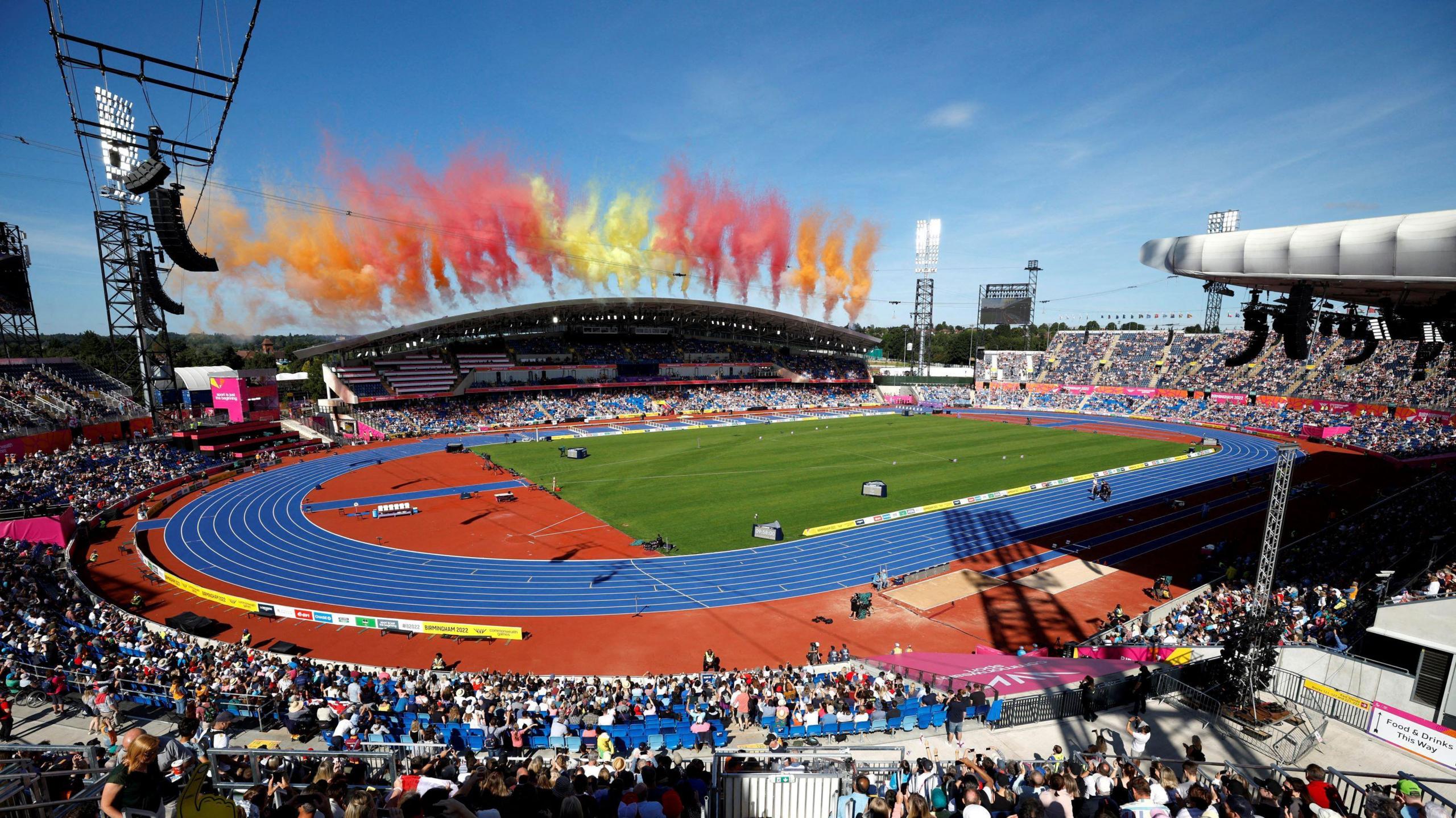 Smoke shot into the air above the Alexander Stadium with a blue track and a grass inner. The smoke is a range of colours at the back of the stadium with an athletics track in the centre and people in the stands.