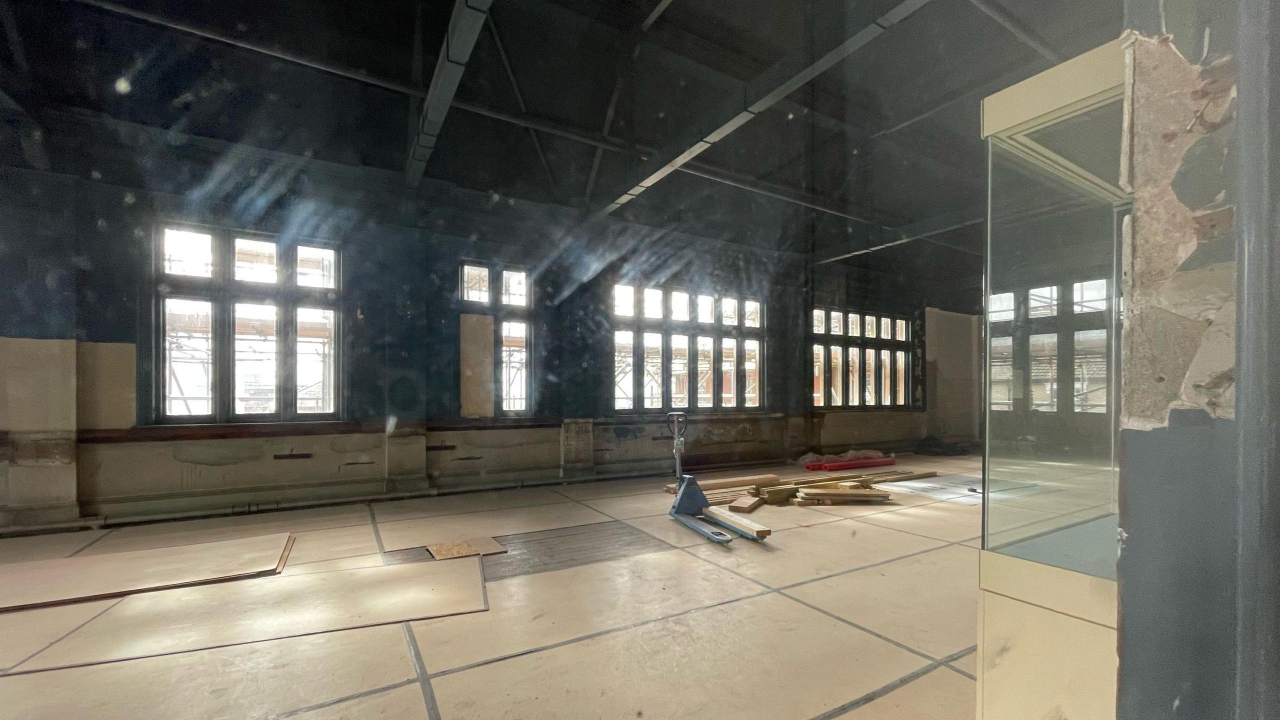 One of the larger rooms in the museum is pictured. Cardboard covers the flood with several of the cardboard sheets out of place waiting to be fitting, Windows line one side of the wall while there are wooden beams toward the ceiling. An empty display case rests against another wall.