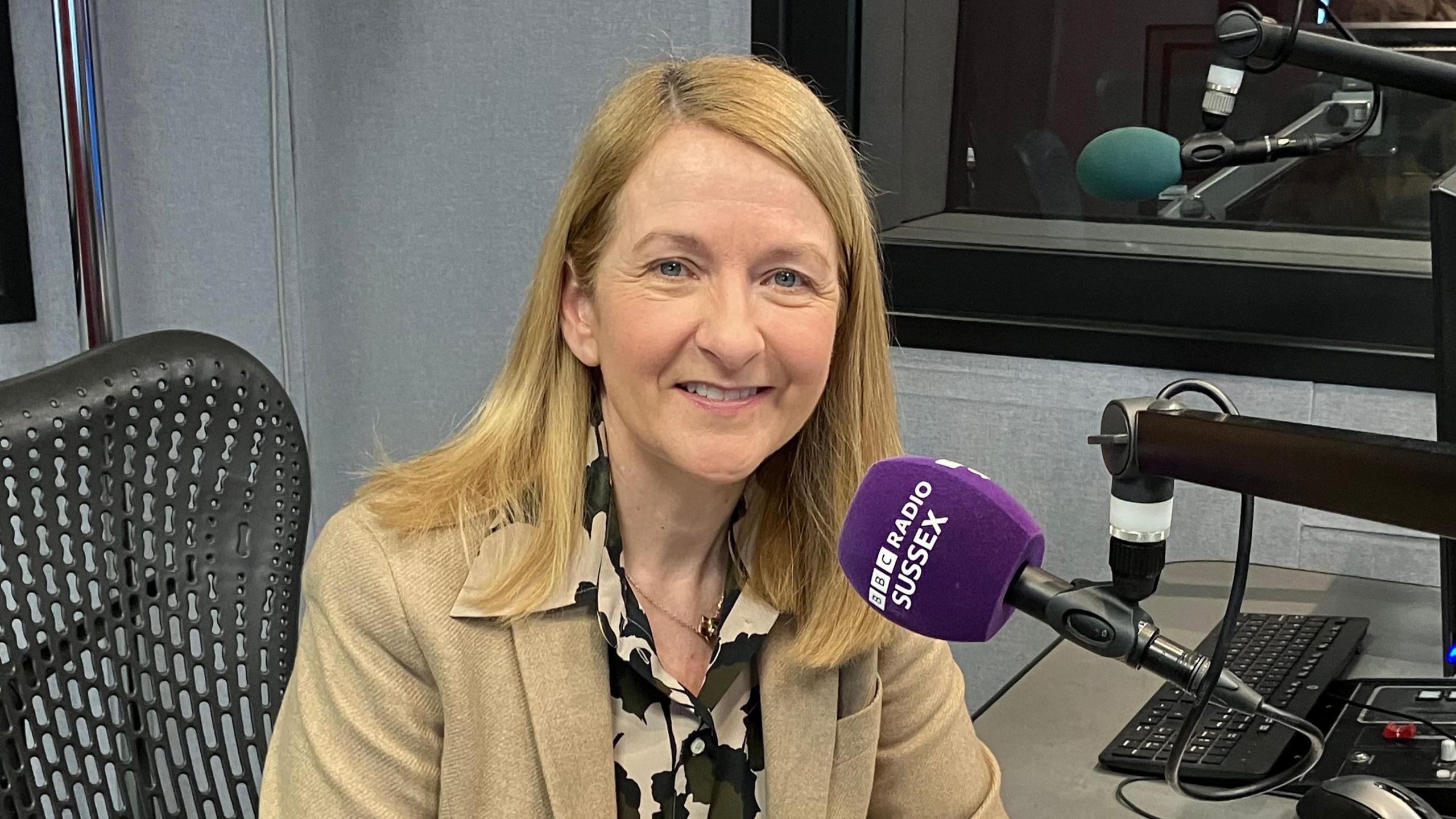 A blonde woman in a brown blazer and black and brown floral shirt. She is sitting behind a microphone with BBC Radio Sussex on it.