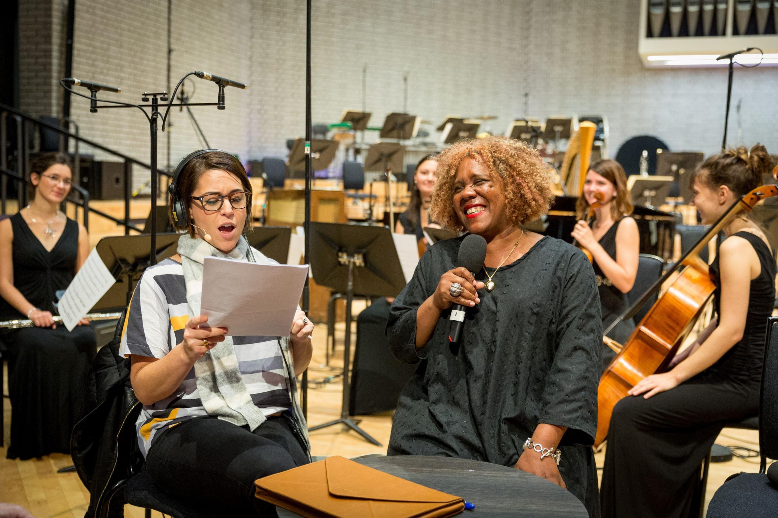 Errollyn Wallen with music students who have a variety of instruments including a cello and a flute. Errollyn is holding a microphone and wearing a black top. A woman next to her is wearing headphones and appears to be singing as she looks at a piece of paper.