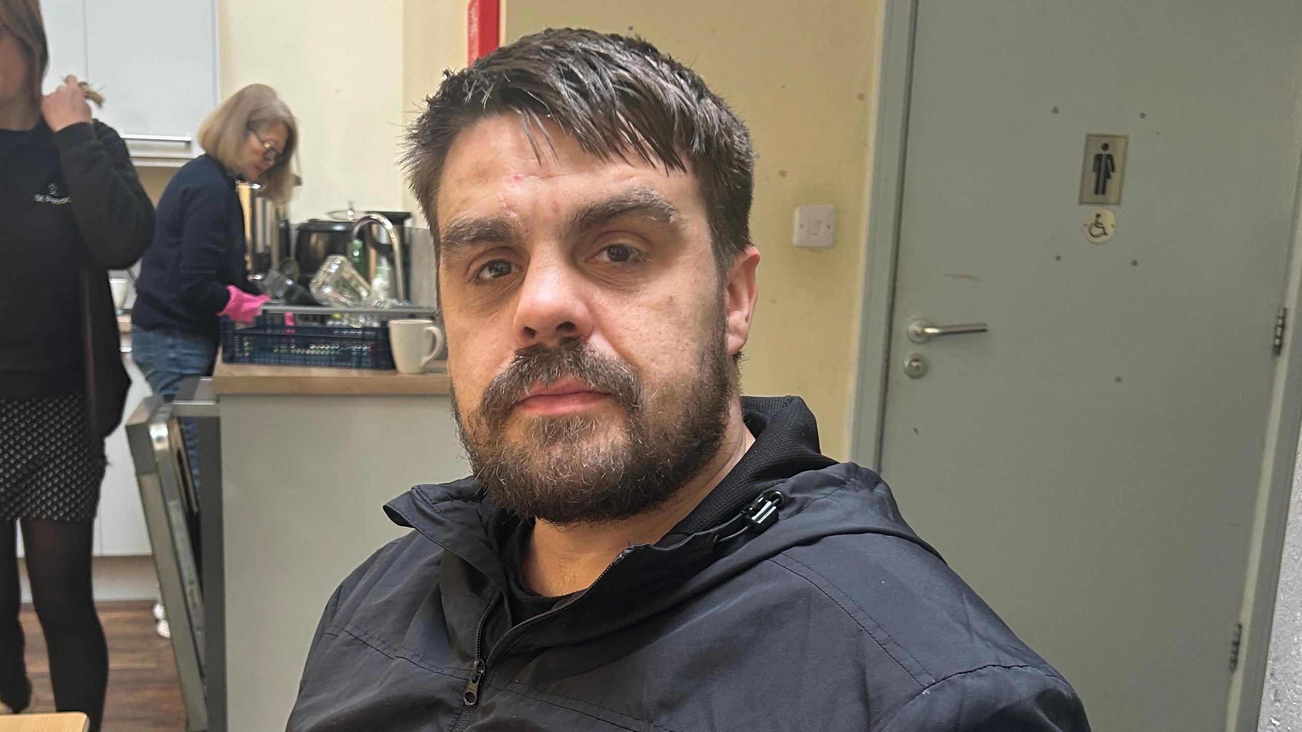 A man with dark brown hair and a beard wearing a black jacket