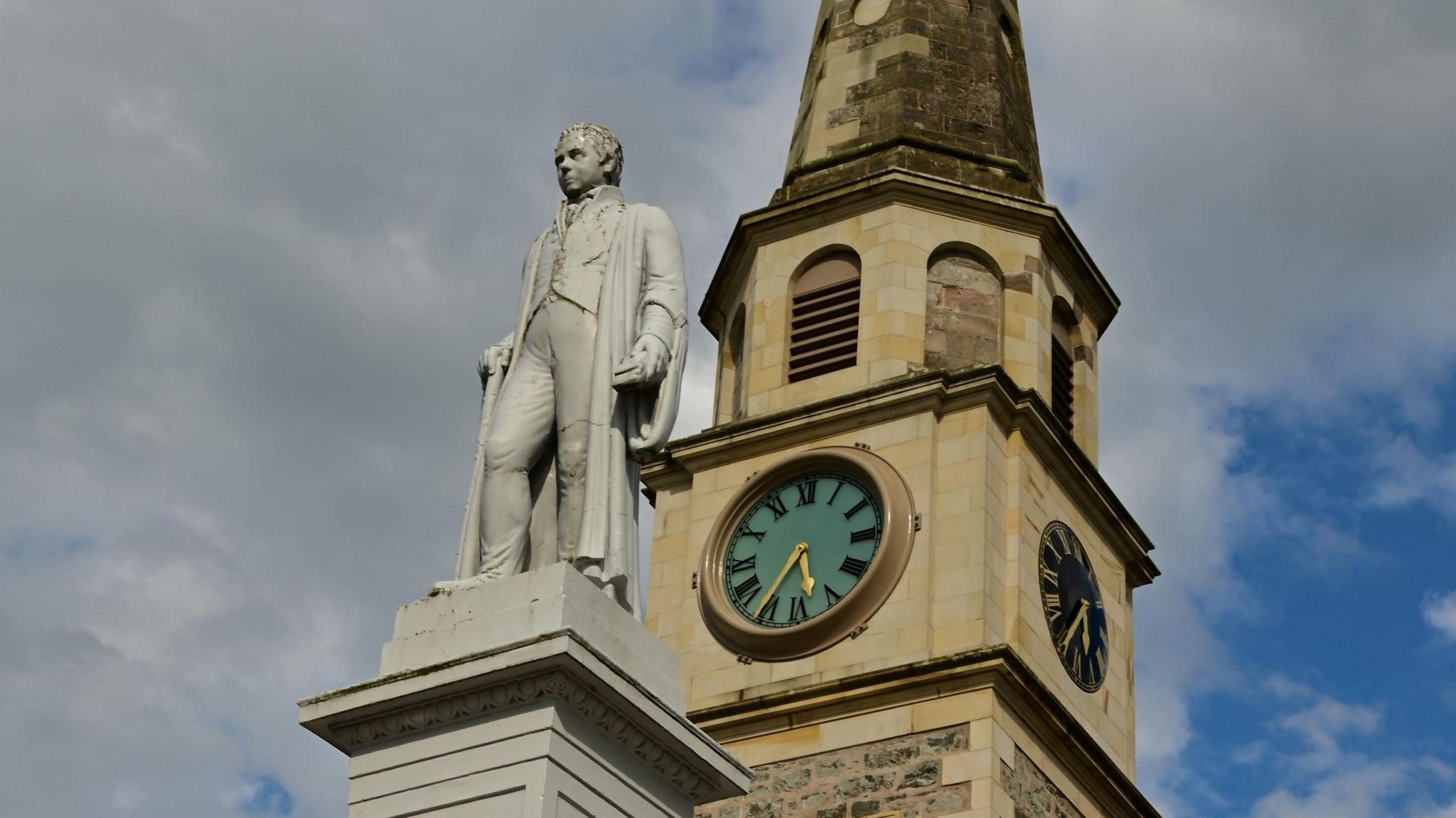 The statue and plinth of Sir Walter Scott is grey in colour. The figure of Sir Walter Scott is wearing a waistcoat, breeches and a gown. The statue stands outside the courthouse in Selkirk. The stone-built courthouse has a clock tower.