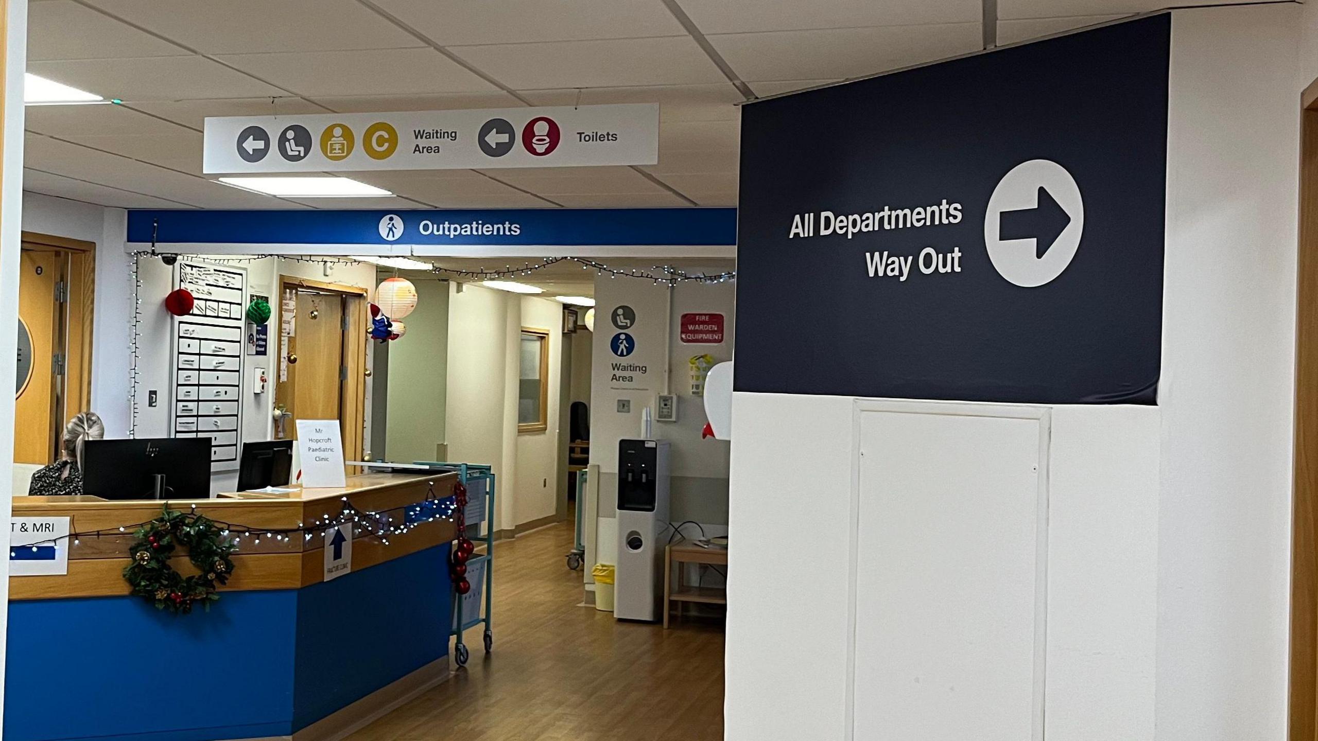 A hospital reception. The desk is blue and has Christmas lights and decorations over it. There is a sign above the desk that reads Outpatients and another to the left side that reads all departments way out. 