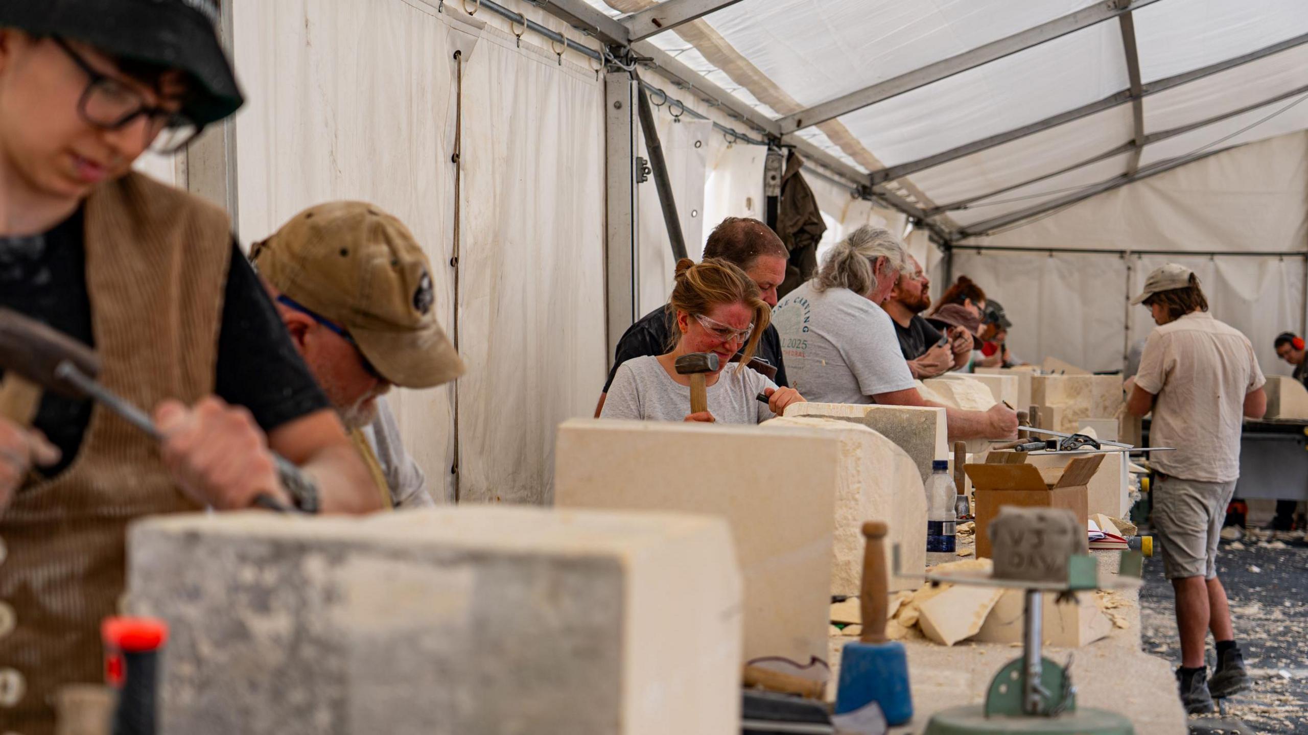 A group of people stand along a table carving stones with hammers under a marquee. There is a lot of dust everywhere.
