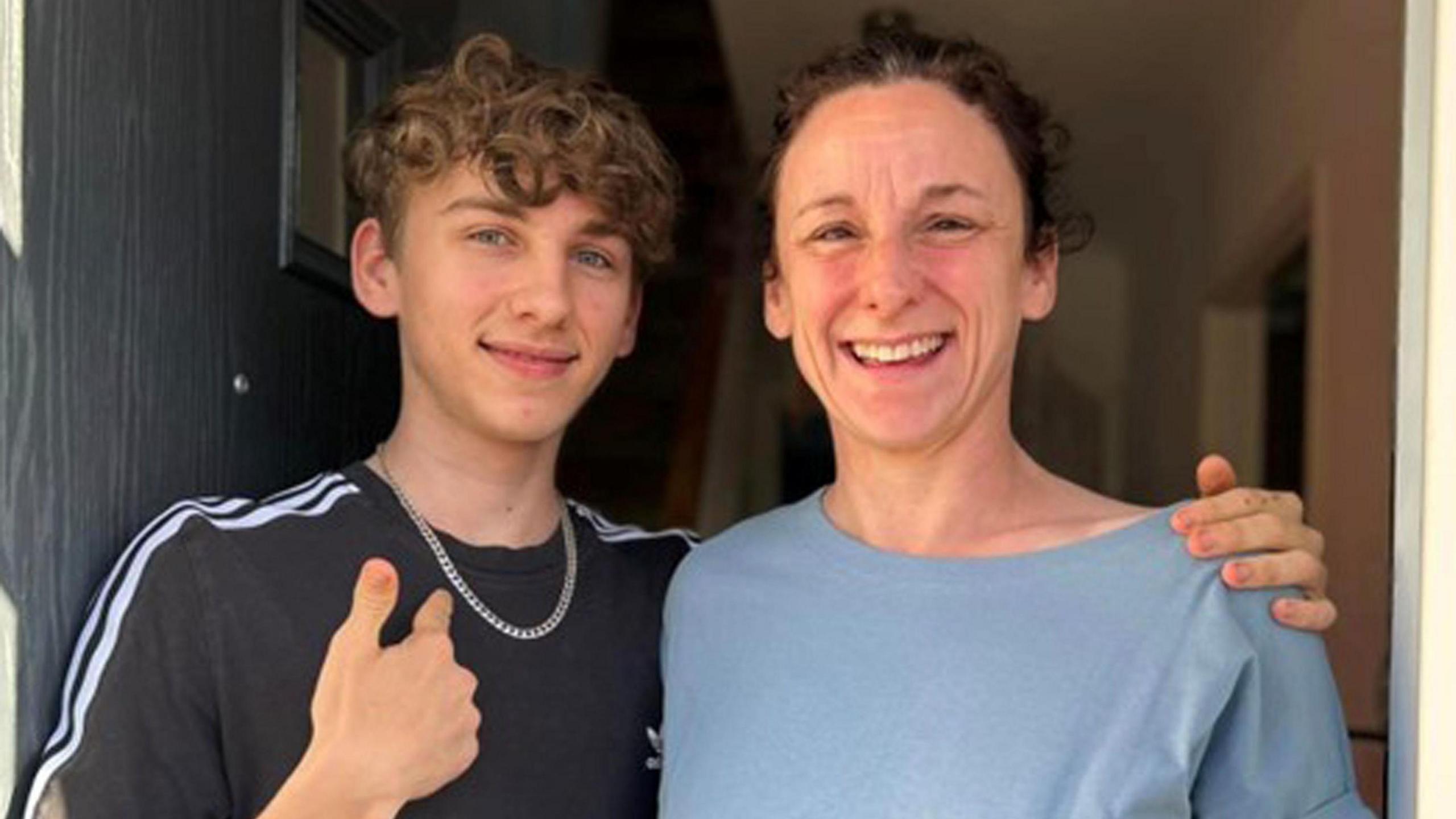 Jamie Holland standing in a doorway with his mother Carrie Holland. Both are smiling at the camera and Jamie is pointing to a silver chain around his neck.