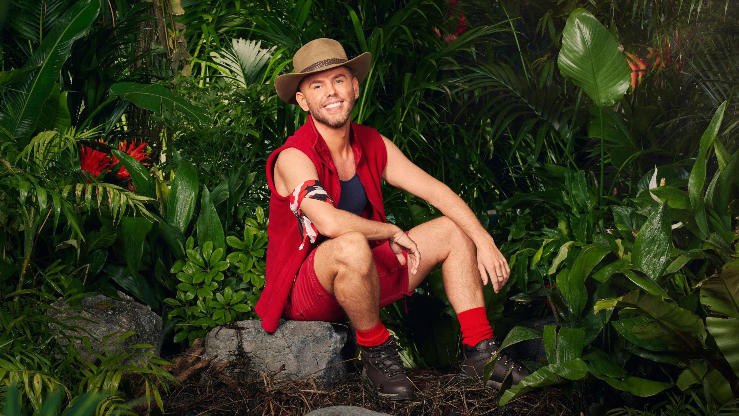 Dean McCullough sits on a rock in his jungle outfit, red socks, shorts and gilet, with a navy vest and earth-toned hat, surrounded by plants 