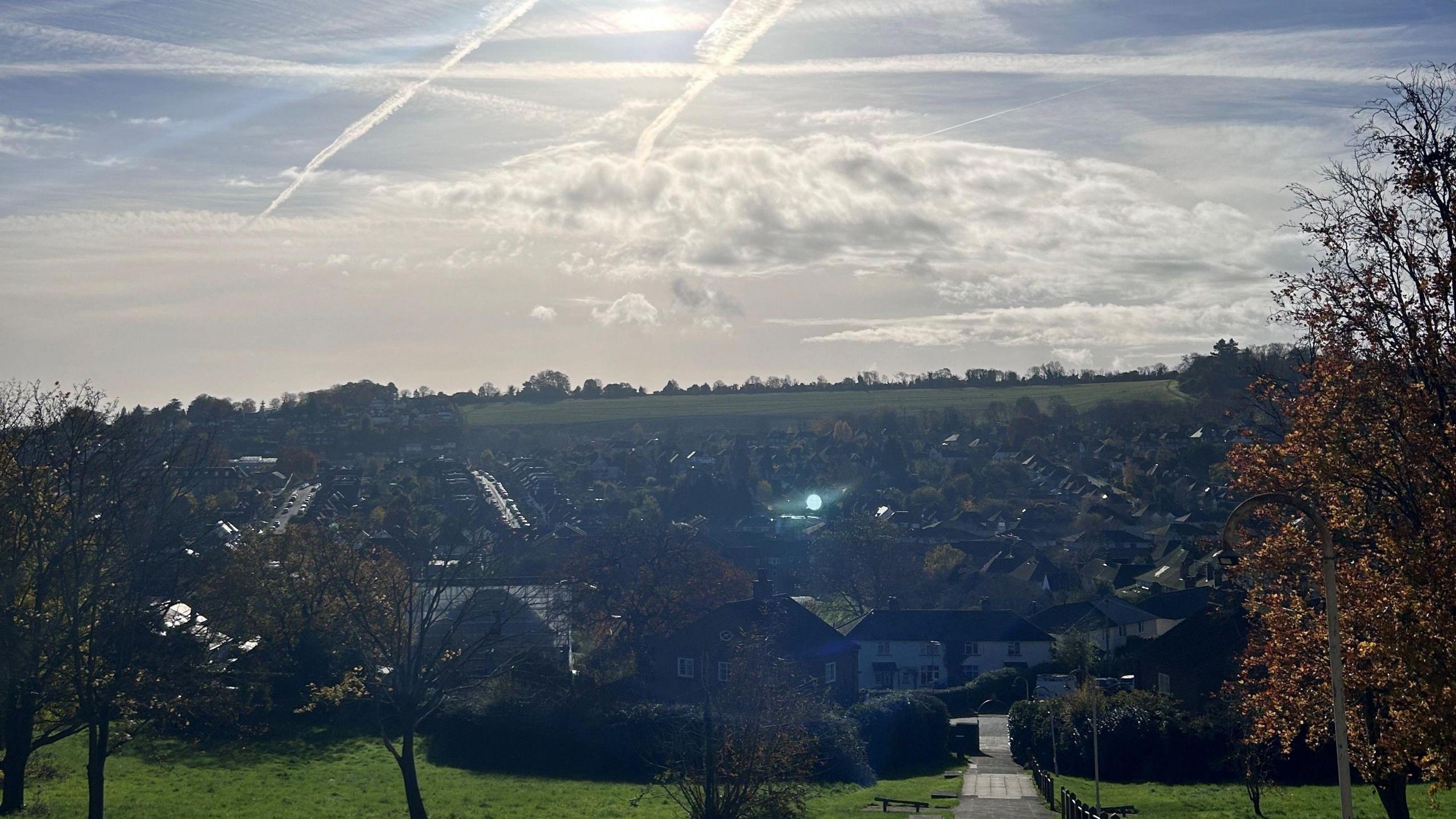 A look down into Guildford, the sun in shining. In the centre of the image is lots of buildings, in the background is fields and trees. There are three trees in the foreground of the image. 