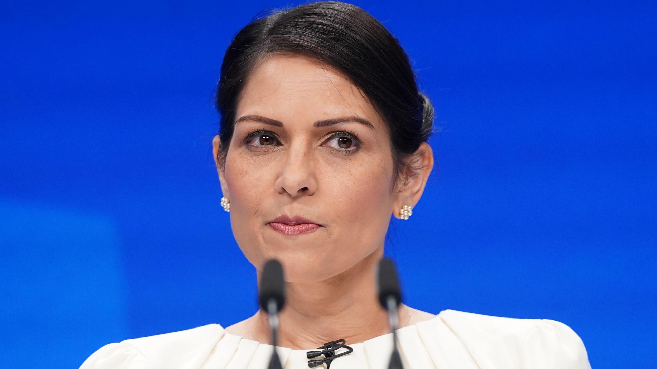 Dame Priti Patel delivering a speech in front of a blue background