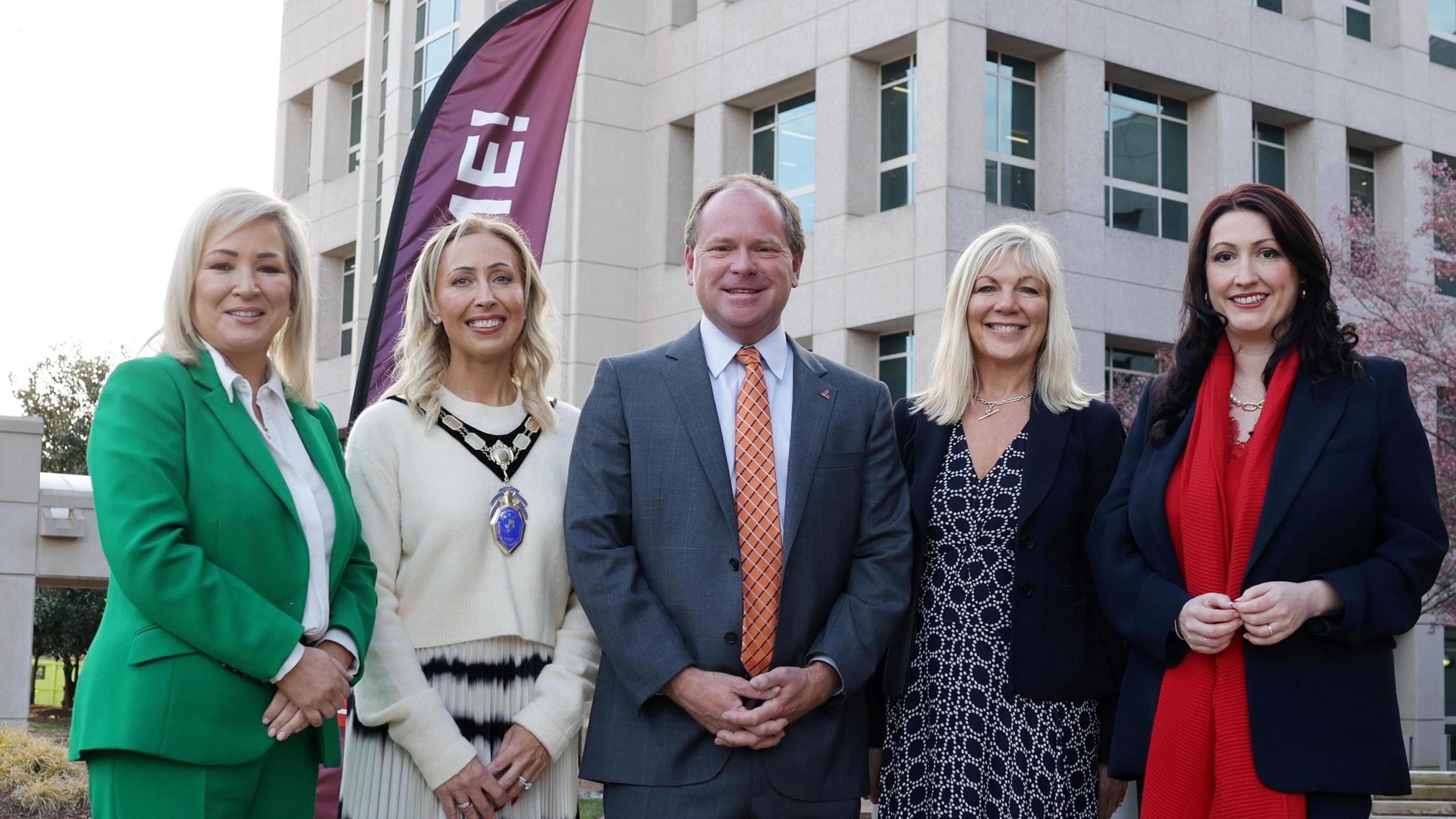 Four women and a man face the camera and are smiling. The man is in the middle of the picture and he is wearing a dark suit and orange tie. MIchelle O'Neill is to the left of the picture - she's wearing an emerald green trouser suit and white blouse and has shoulder length blonde hair. Beside her is a woman with long blonde curly hair who is wearing a cream coloured top and skirt with a black pattern at the top. She also wears a large medal around her neck, to signify that she's from the NI chamber. On the far right is Emma Little Pengelly wearing a dark jacket and red scarf. She has long dark hair. Beside her is another blonde haired woman wearing a dark patterned dress and dark jacket.