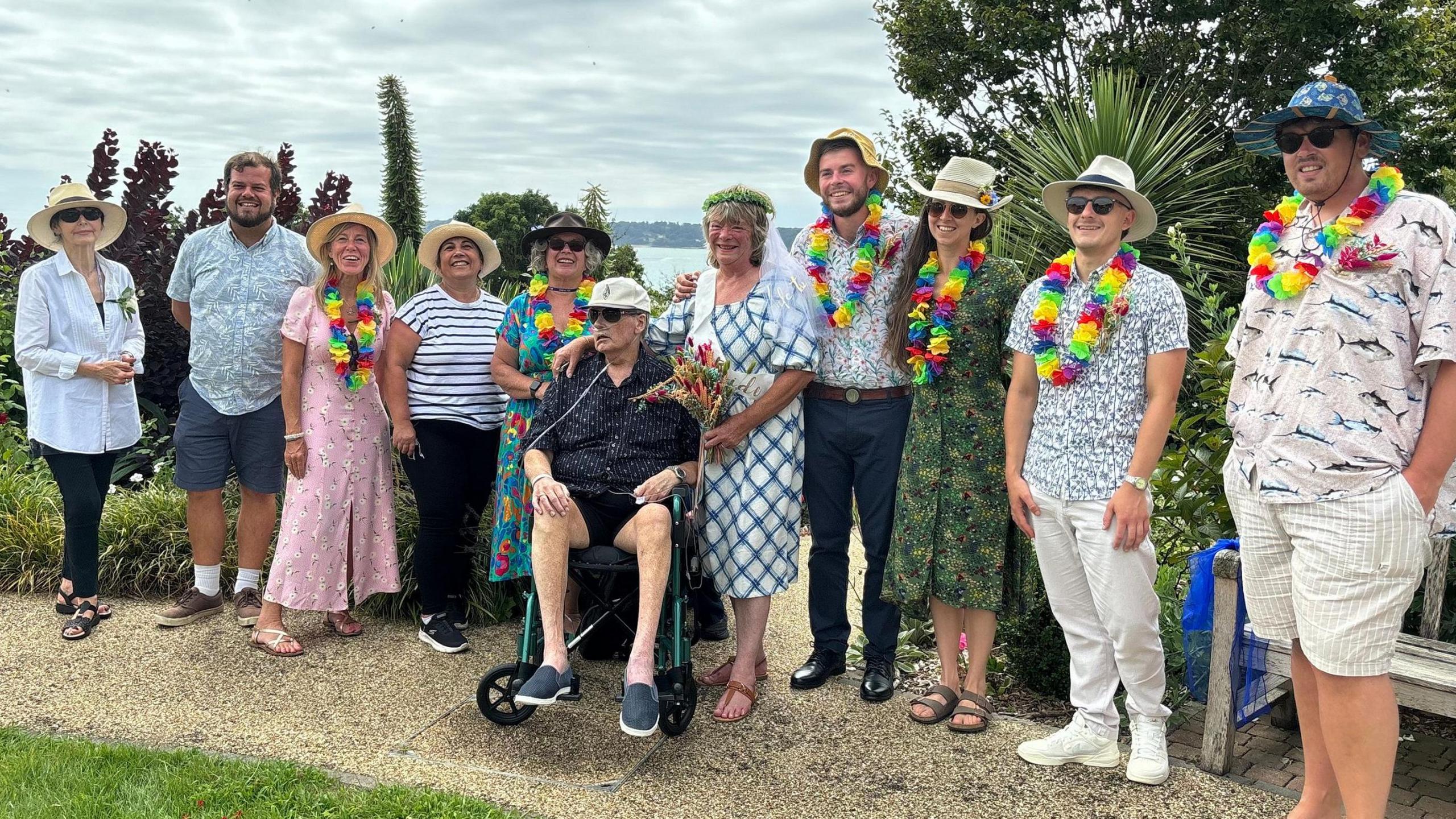 David is in a wheelchair with a breathing tube attached to his nose and Alison has her arm around him. People stand next to them and they're all smiling after the wedding service. Many are wearing bright clothes, hats and sunglasses