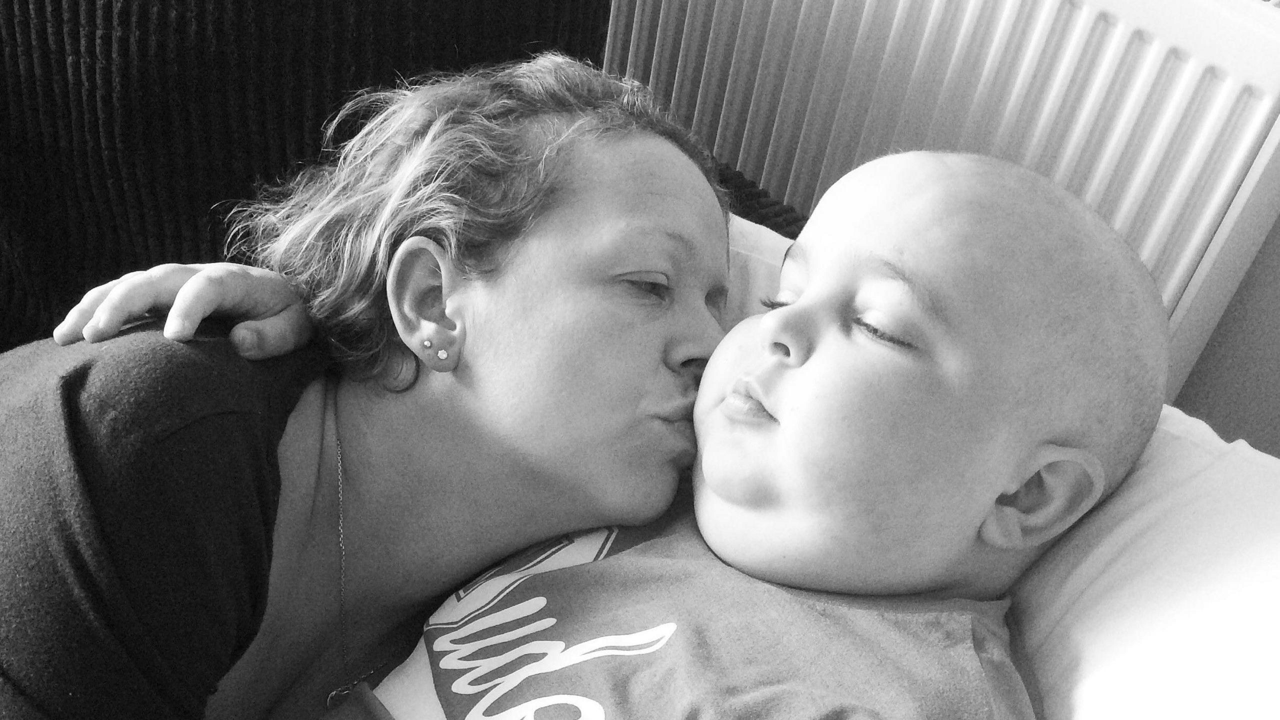 A black and white image of a woman hugging and kissing a young boy who is bald and unwell. Both of them are lying on a bed cuddling each other.