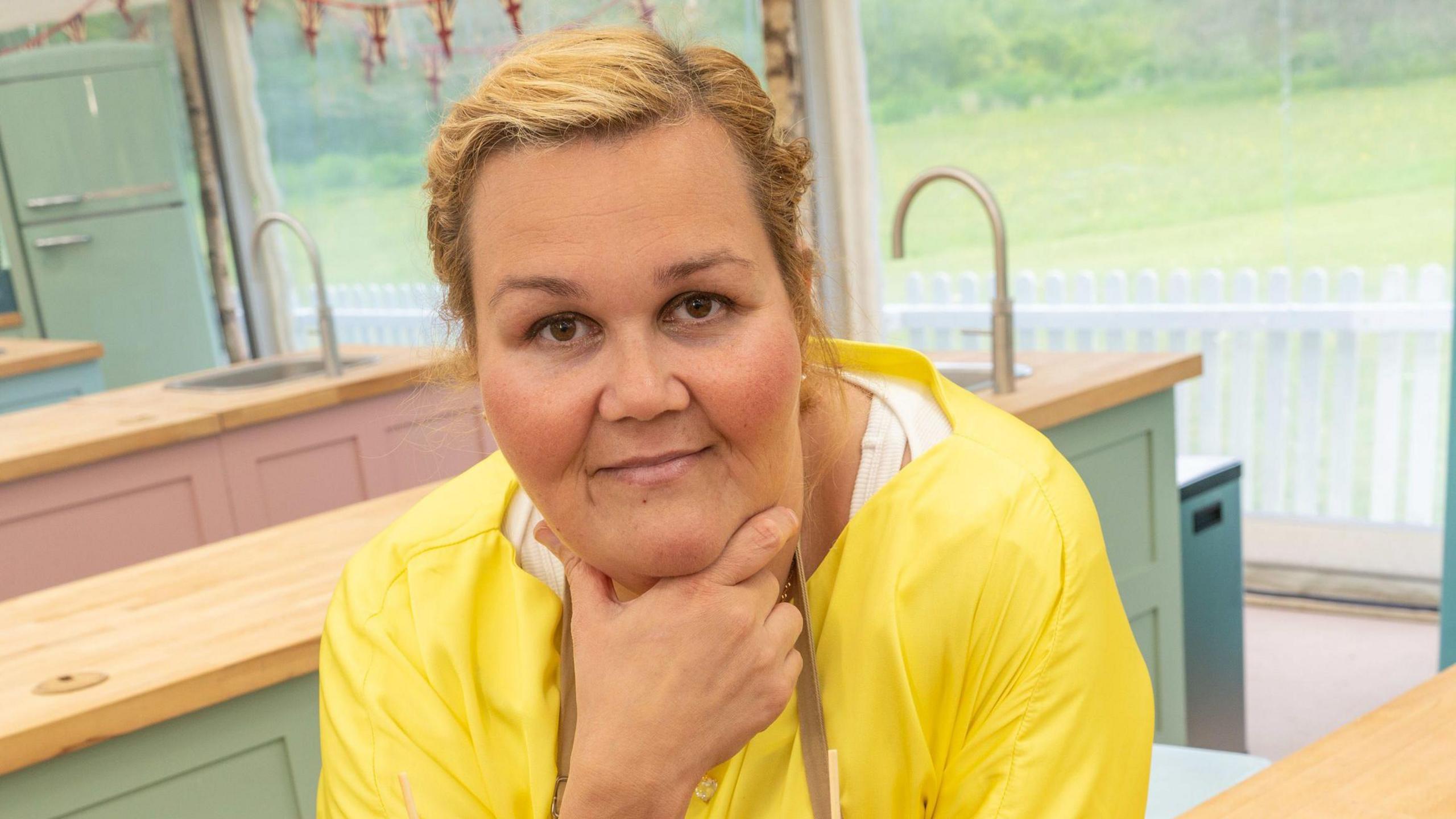 Nelly Ghaffar posing for the camera in the Bake Off studio tent. She has blonde hair and is wearing a yellow shirt