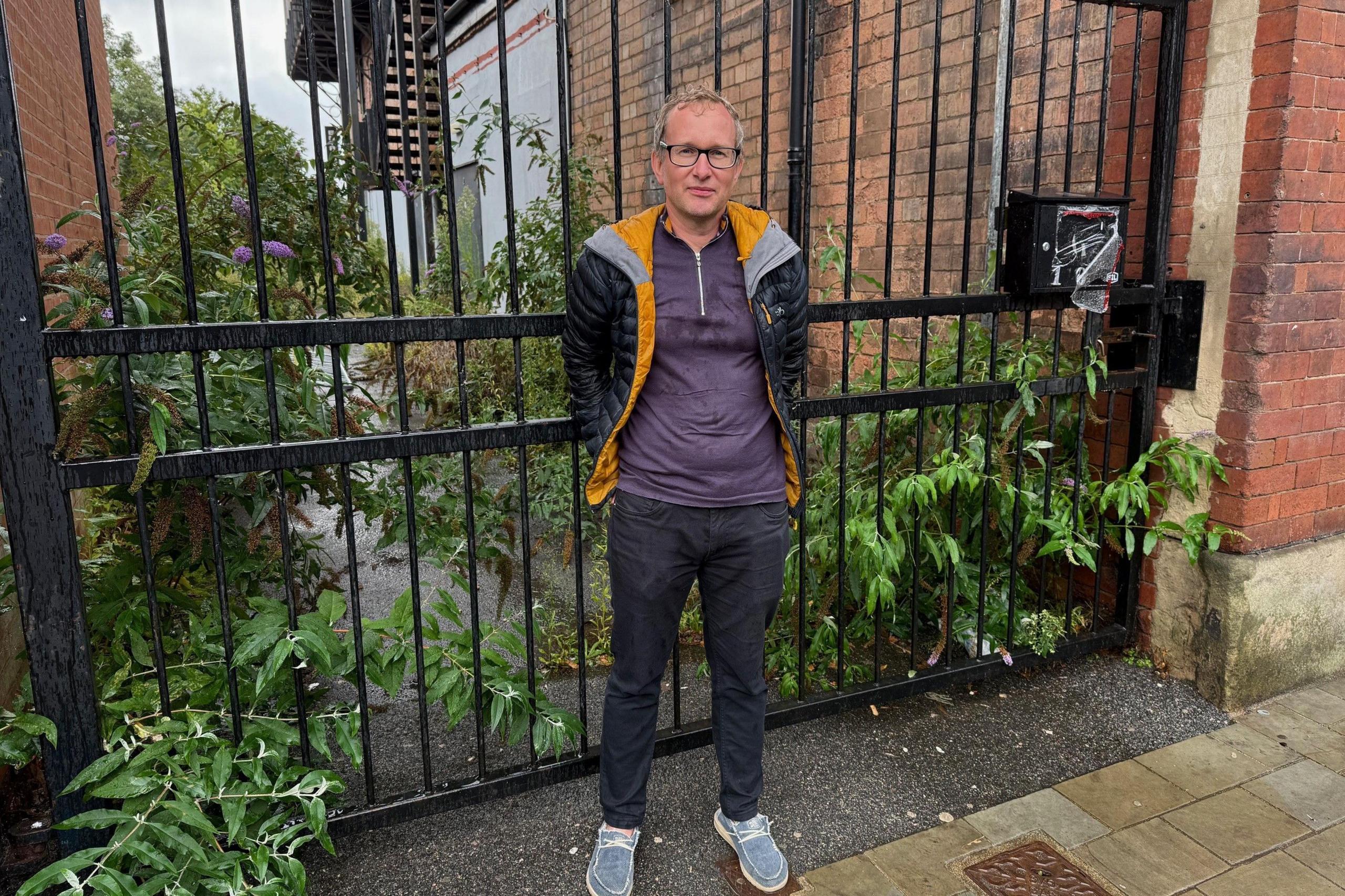 Oliver Astley stood in front of some black gates with weeds in the background