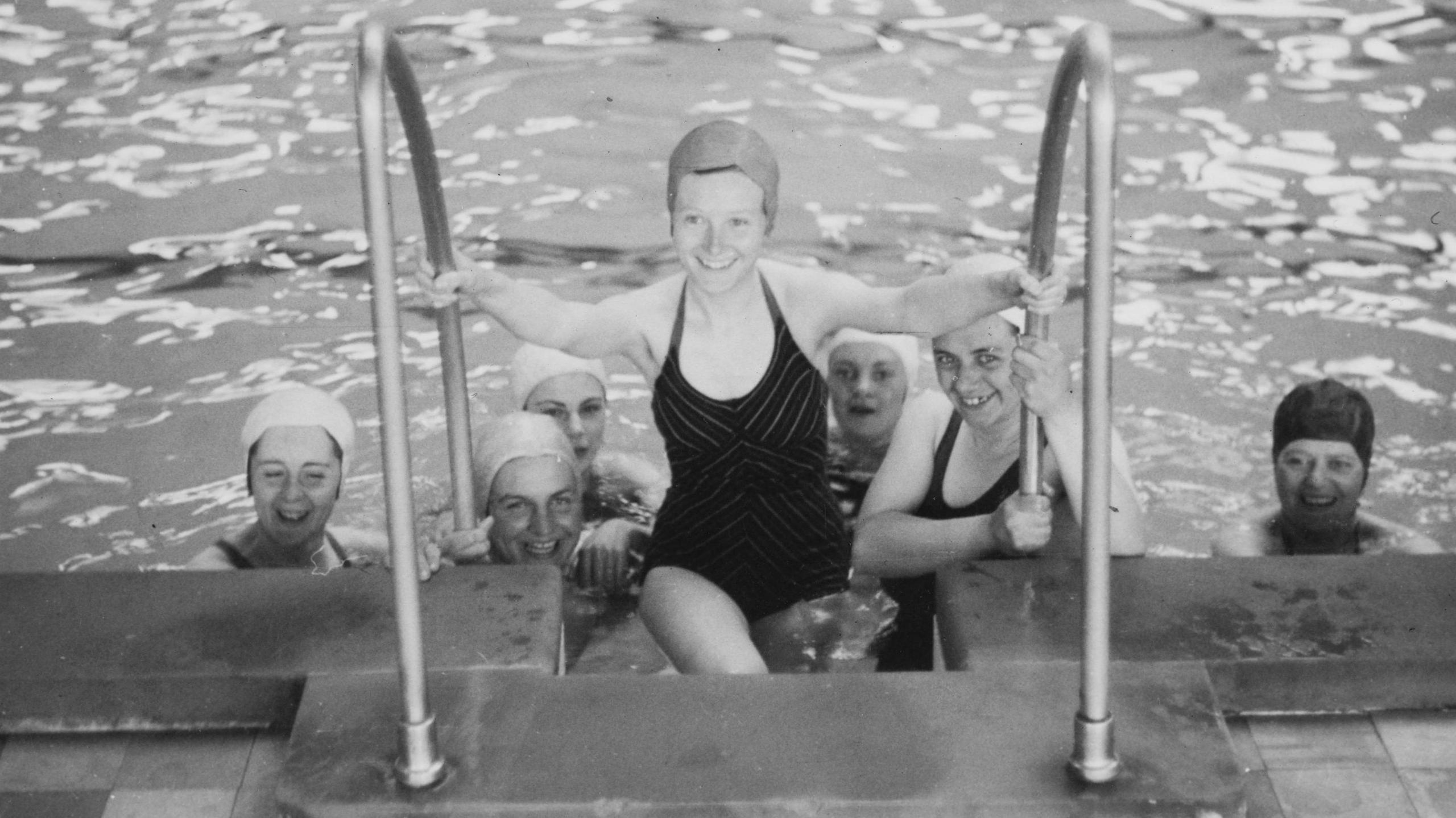 Seven women can be seen in a swimming pool wearing swimming costumes and swimming caps. One of them is walking out of the pool towards the camera.
