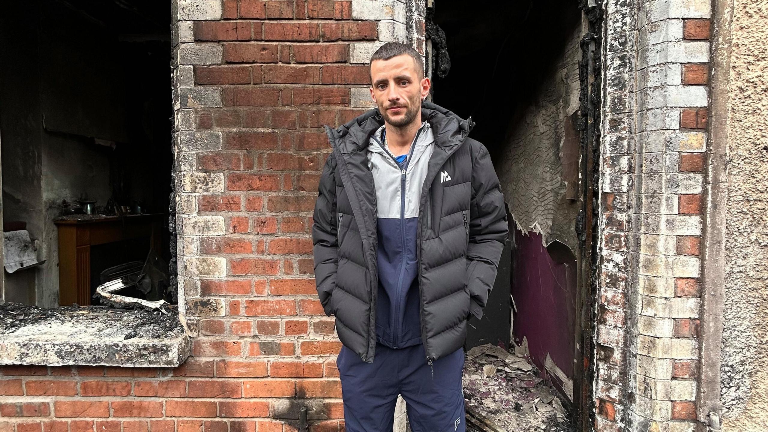 A man with brown hair and a black puffer coat stands outside his home 