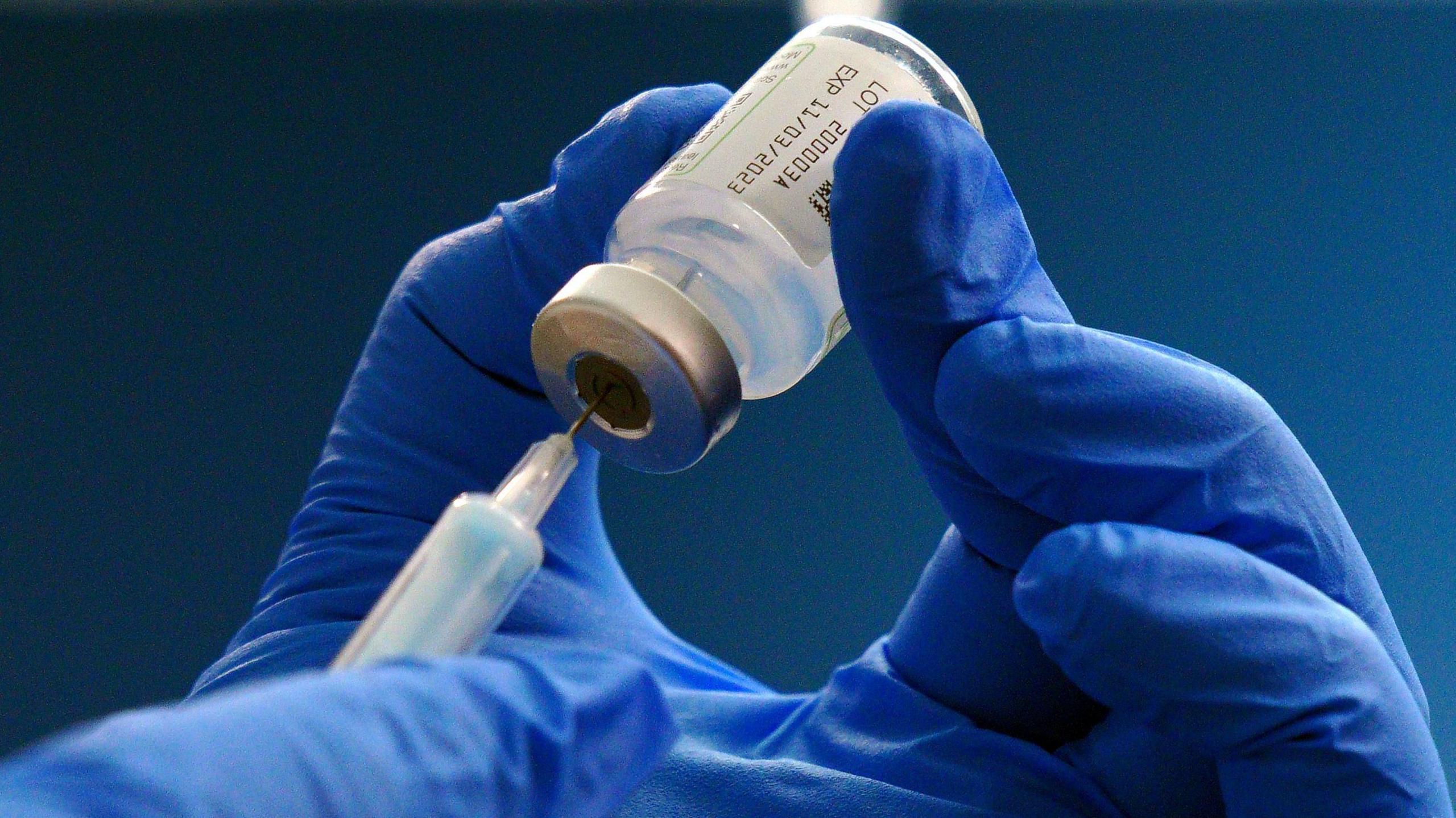 A covid vaccination bottle is held in the air while someone in blue rubber gloves holds up a needle to the vial